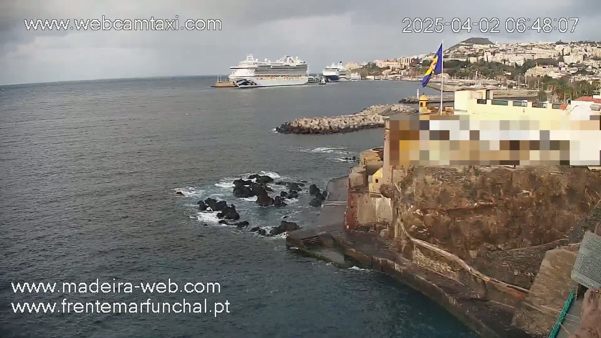 A large cruise ship is docked in a harbor next to a rocky shoreline with a building on top, under an overcast sky.