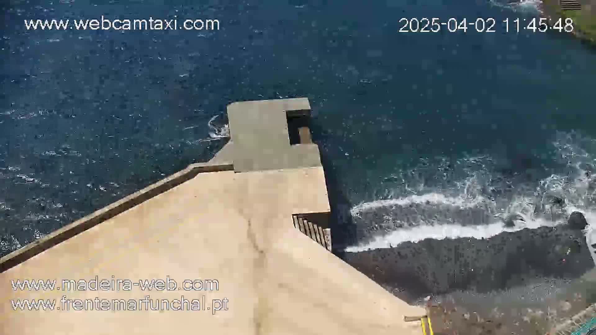 An aerial view shows a light-brown concrete structure extending from a rocky shoreline into a dark-blue ocean with small waves crashing against the rocks.