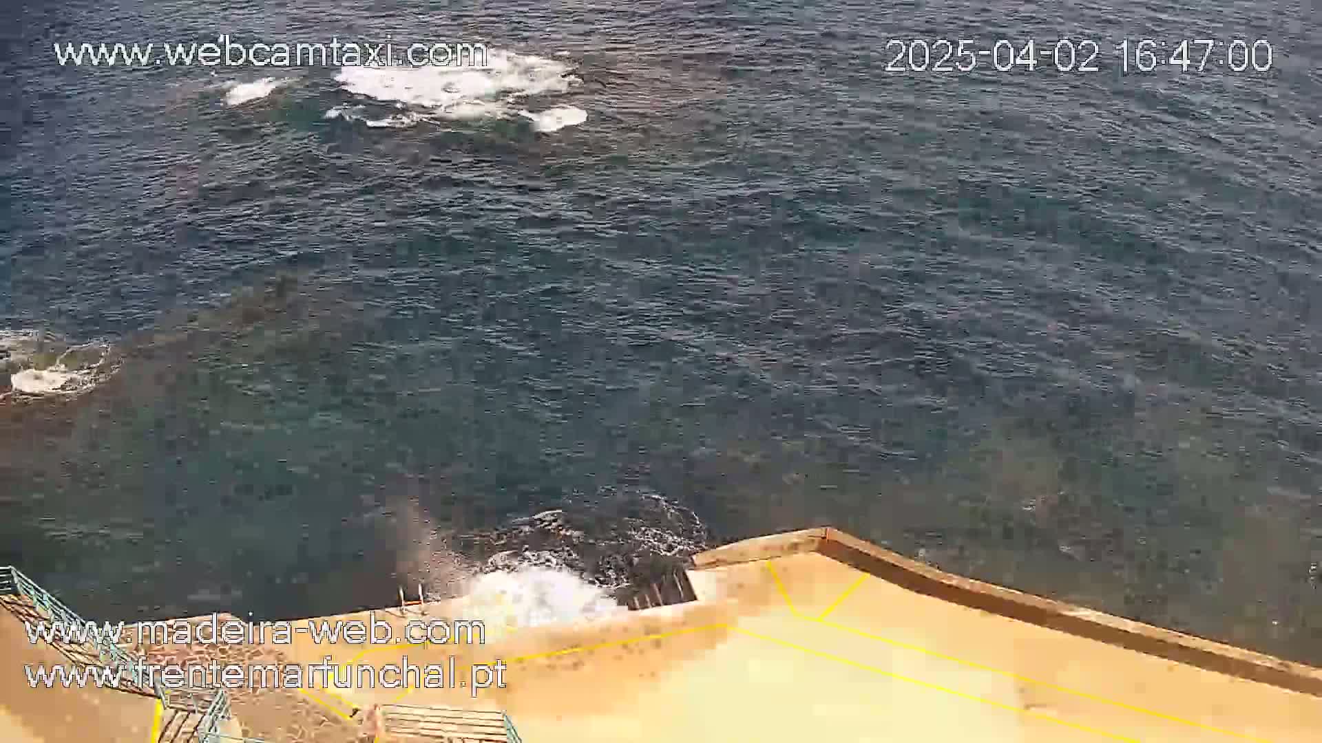 A high-angle view shows dark-blue ocean water with small waves lapping against a tan concrete platform with stairs leading down to the water.