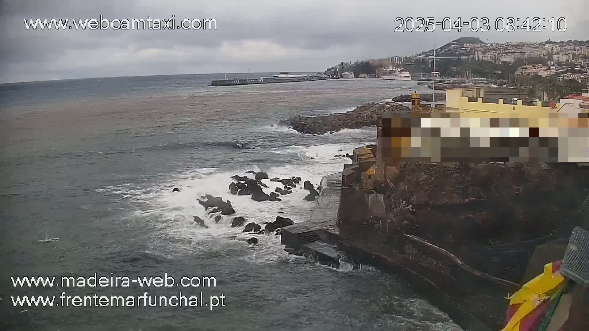 A rocky shoreline with crashing waves under an overcast sky shows a harbor with a cruise ship in the distance.