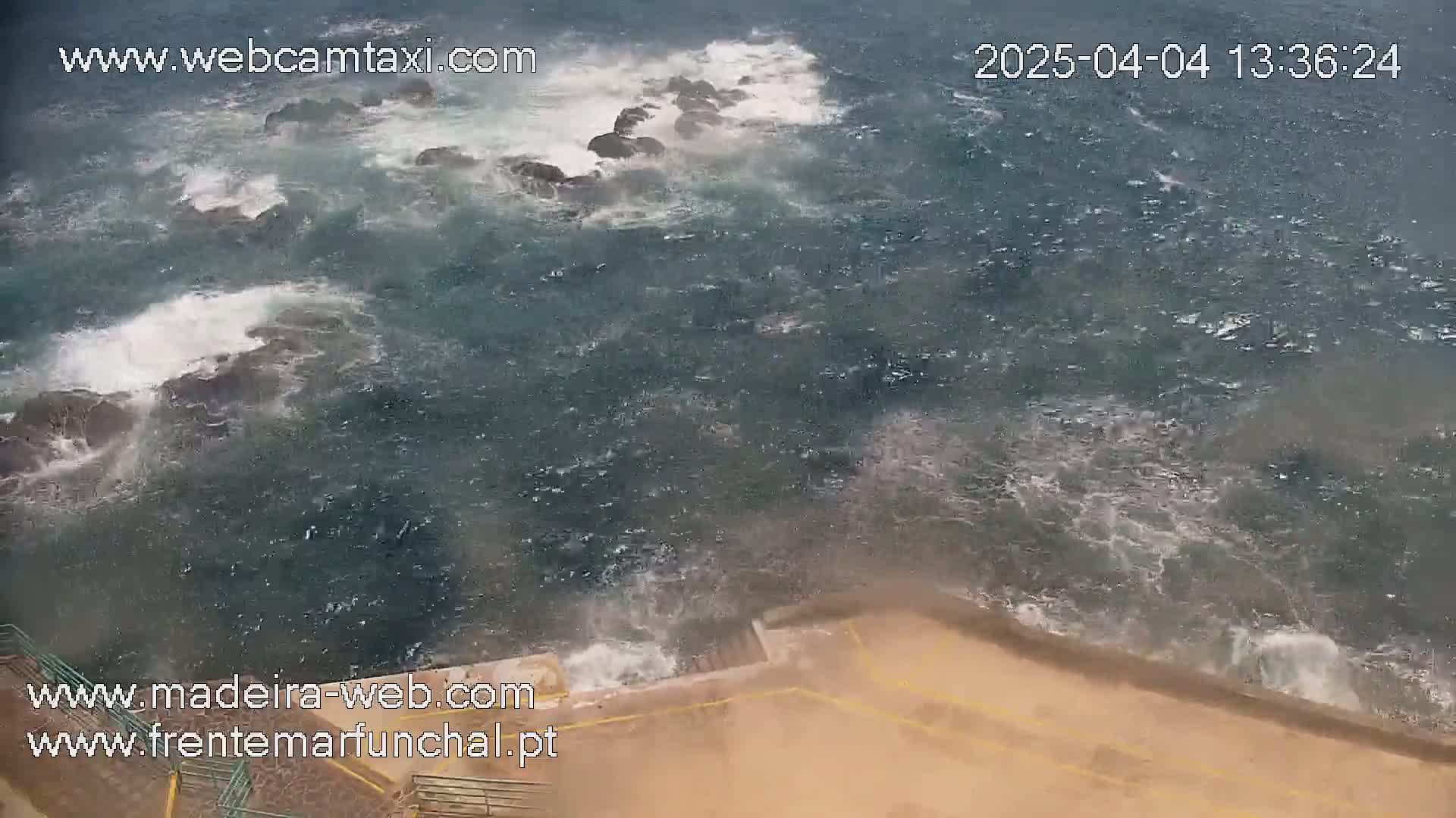 Rough ocean waves crash against dark rocks near a tan, sloped seawall during a windy, possibly rainy day.