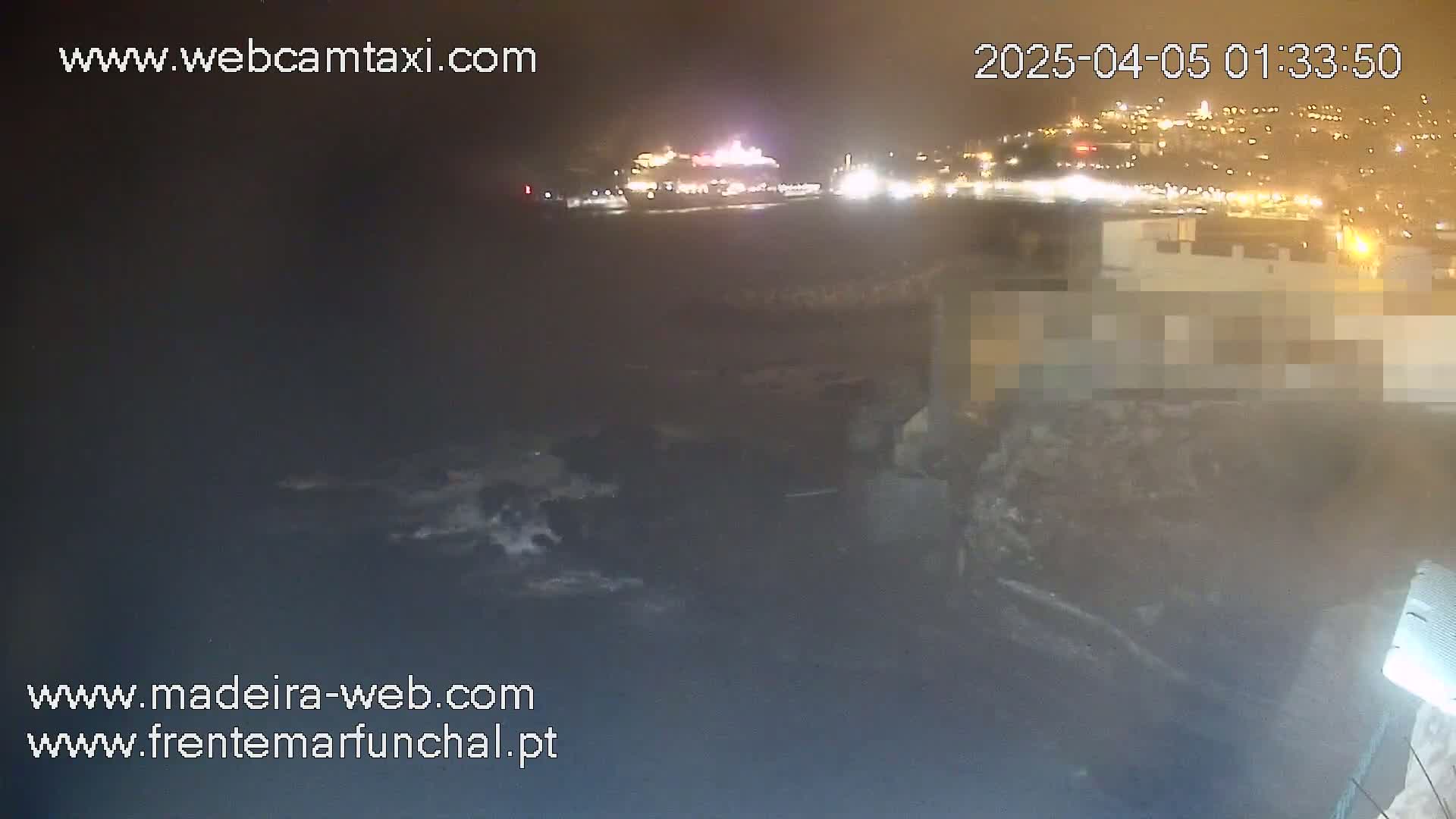 A nighttime, low-light view of a rocky shoreline with crashing waves, a distant city illuminated with lights, and a large vessel visible in the water.