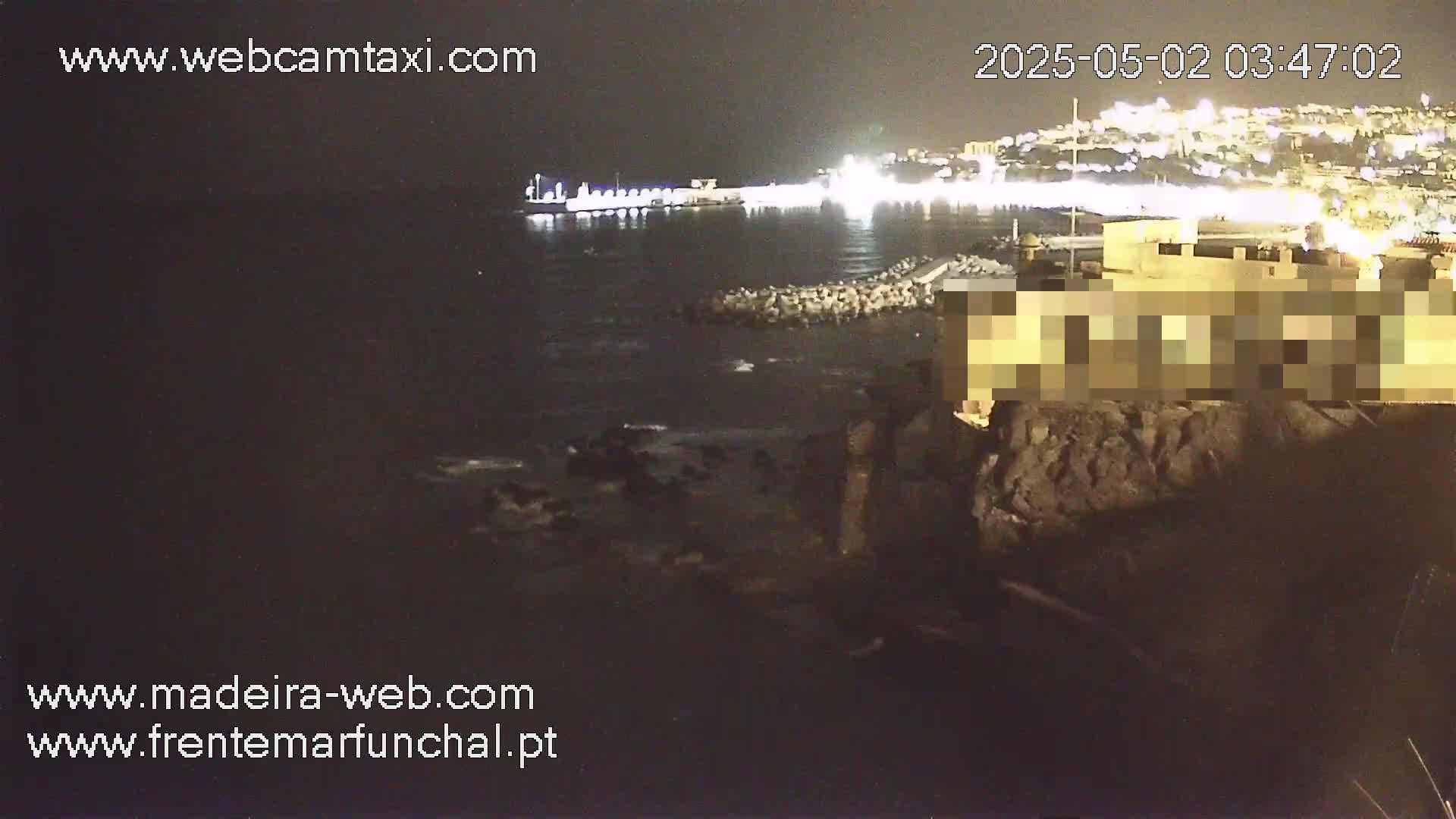 A nighttime coastal scene shows dark water lapping against a rocky shoreline near a brightly lit city.