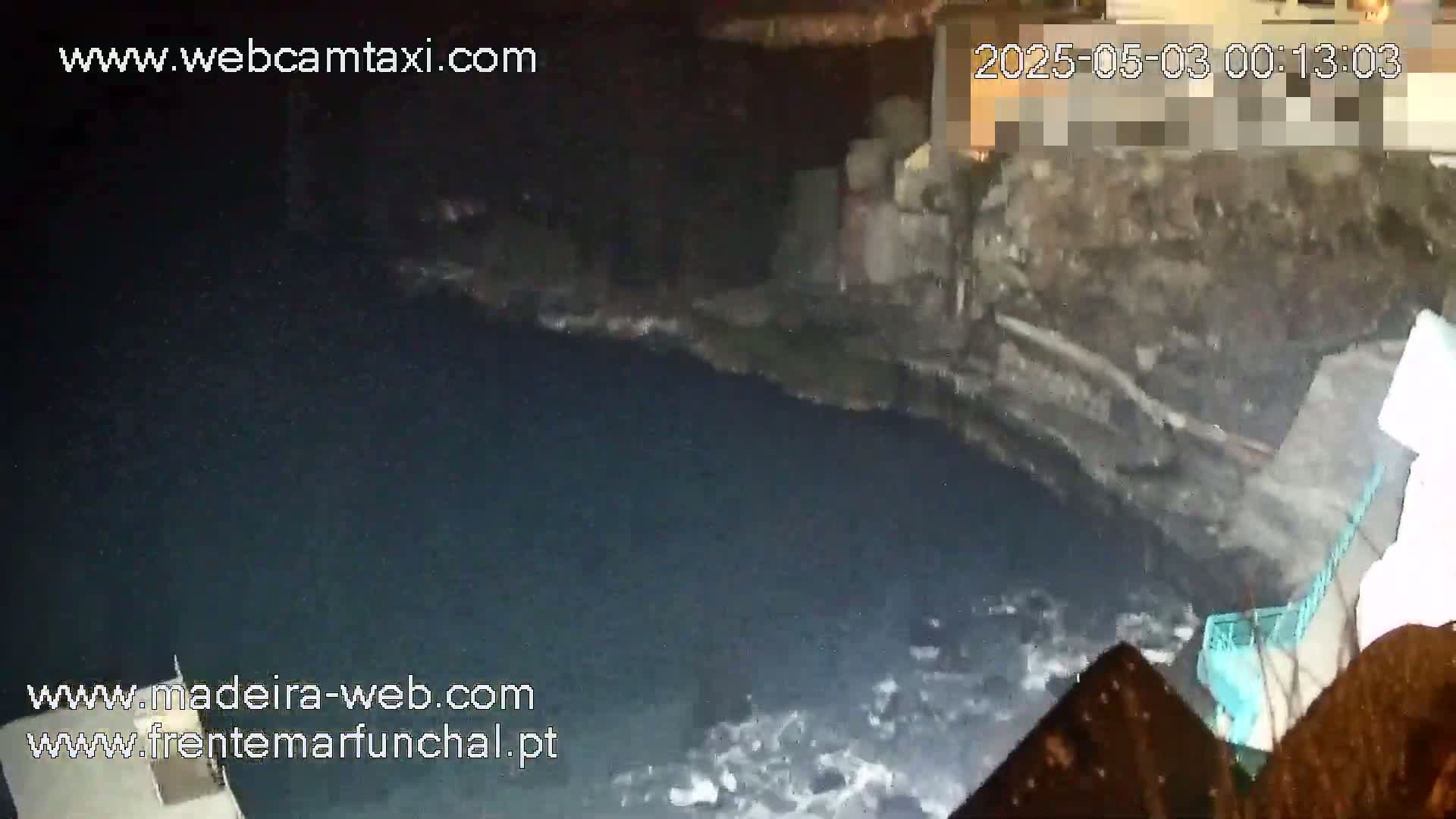 A nighttime, low-light view shows dark, possibly rocky, terrain sloping down to a body of dark water with some small waves or debris near the shore.