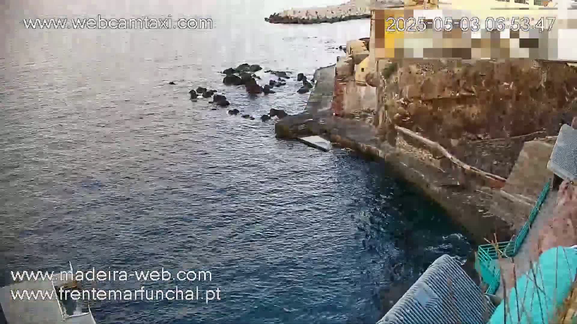Dark, calm water laps against a rocky shoreline near a stone building on an overcast day.