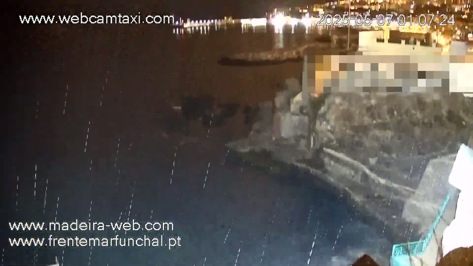 A nighttime, rain-sprinkled waterfront scene shows a dark body of water with a partially visible building and other structures along the shore, illuminated by distant lights.