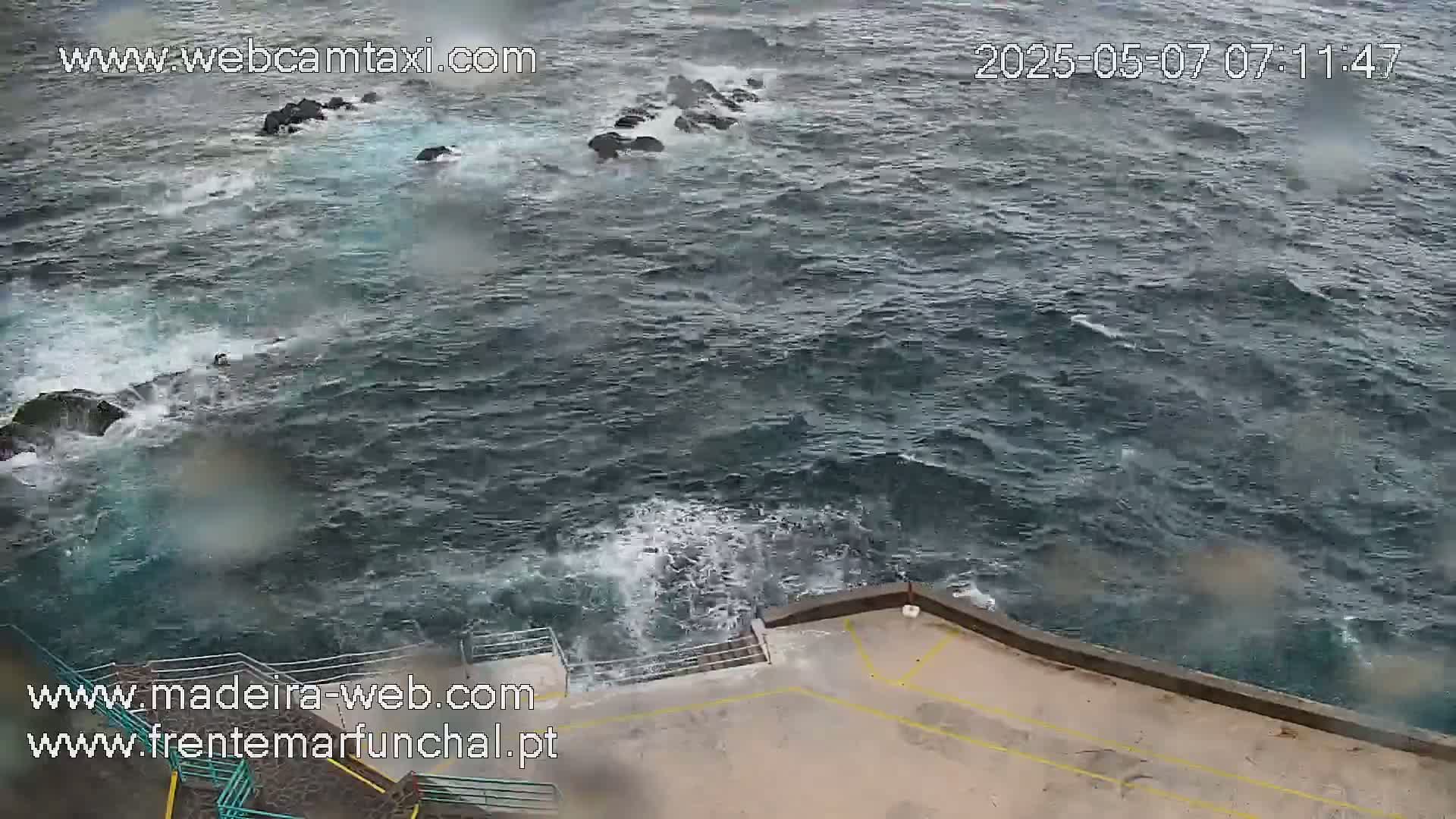 Rough, dark-colored ocean waves crash against dark rocks near a concrete structure with railings.
