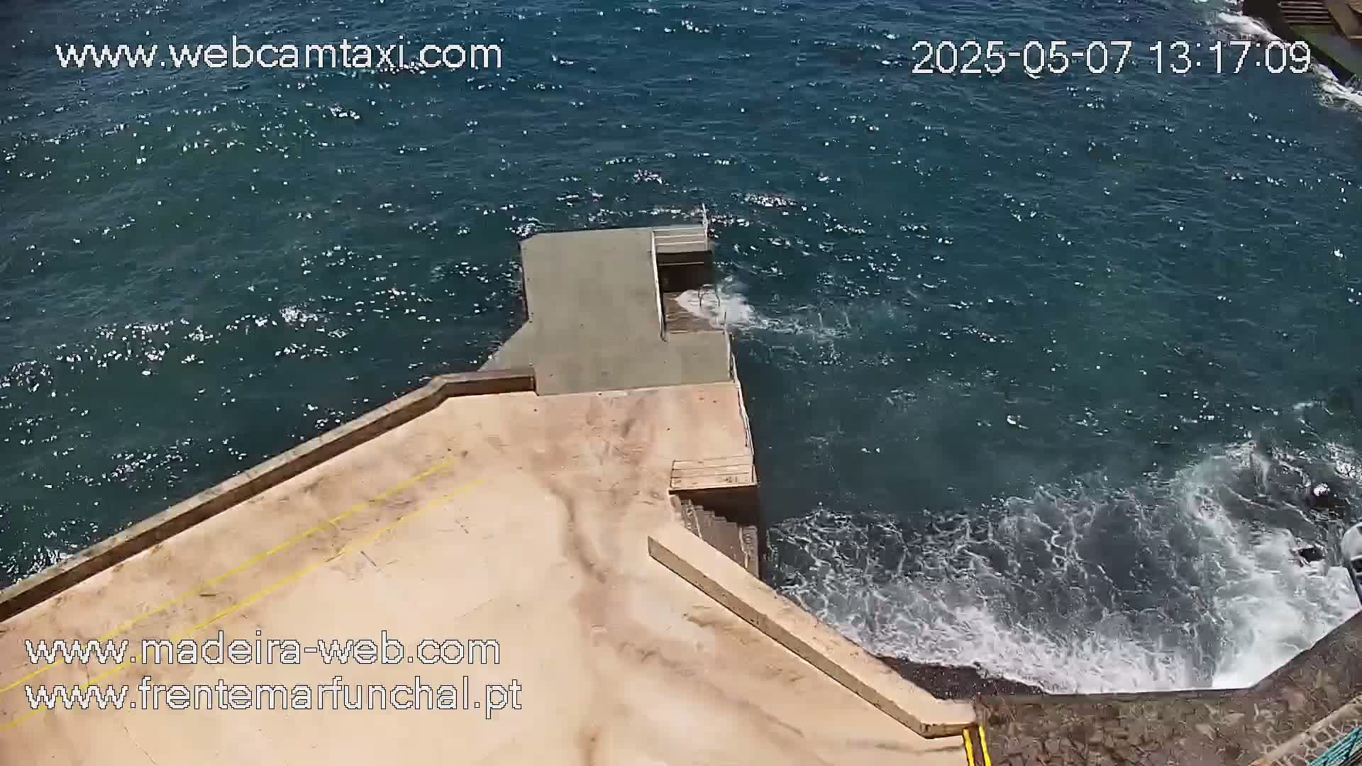 A tan concrete platform extends over a dark-blue ocean with small waves crashing against its edge on a sunny day.