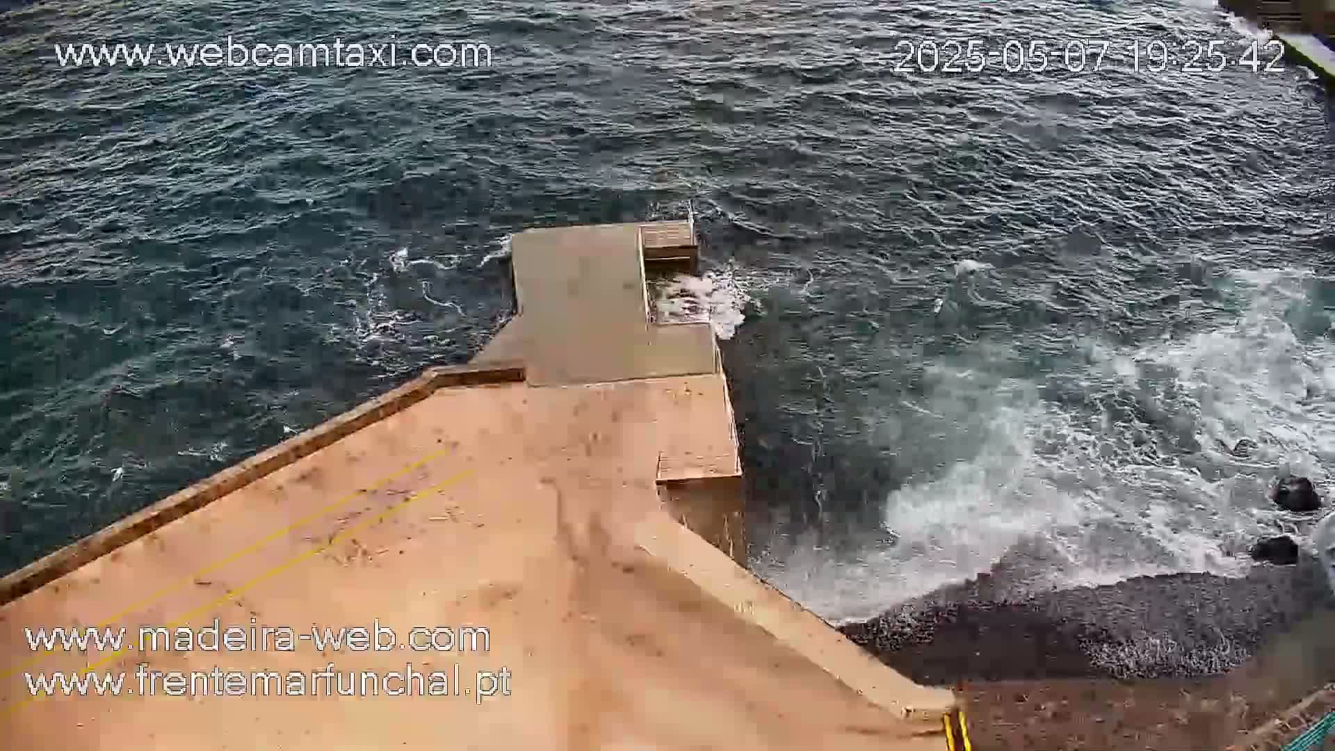 A tan concrete platform extends over dark, choppy ocean water with whitecaps crashing against a rocky shore.