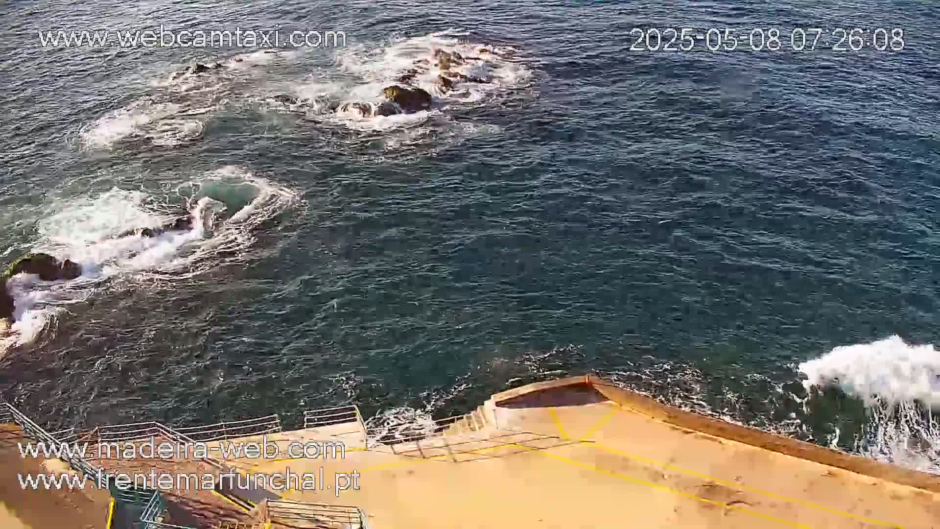 Dark-blue ocean water crashes against dark rocks and a tan concrete wall with steps leading down to the water's edge.