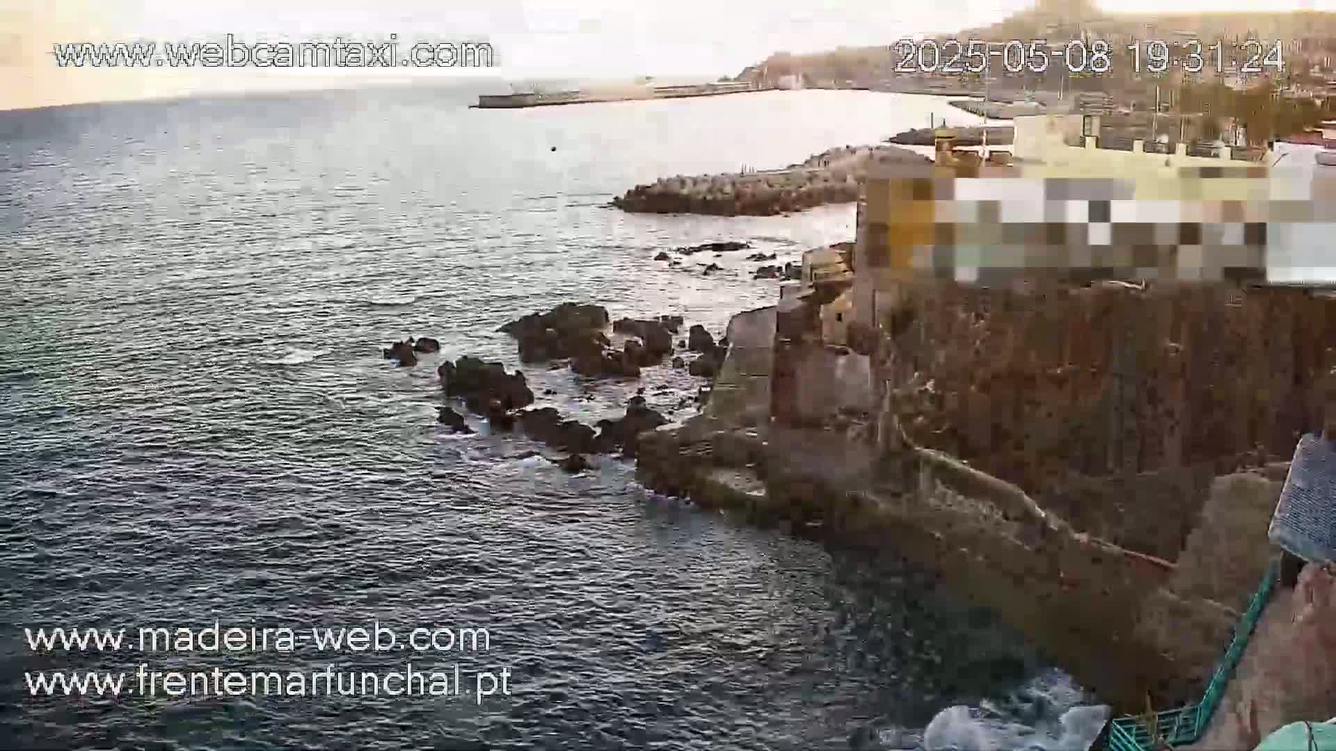 A rocky coastline with buildings is seen from above, overlooking a calm body of water under a partly cloudy sunset sky.