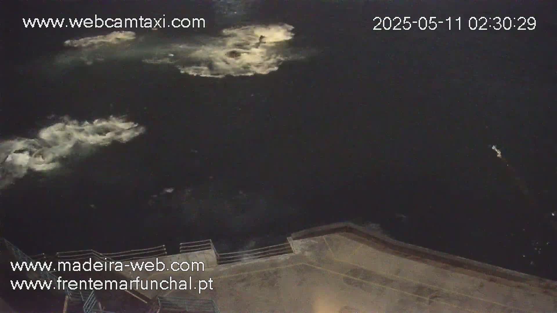 A nighttime, high-angle view shows dark water with faint, white splashes and a section of a light-brown concrete structure.