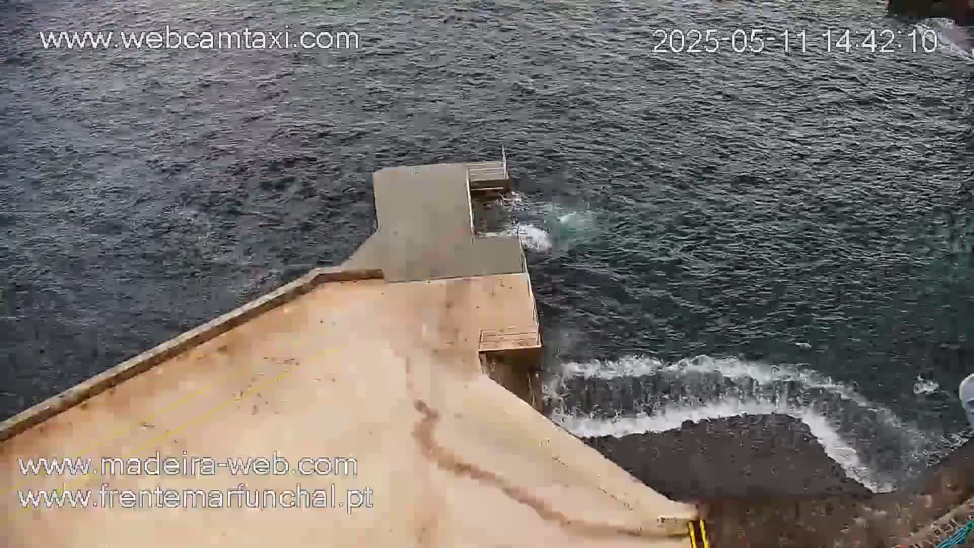 An aerial view shows a small concrete pier extending into a dark, choppy ocean with small waves lapping against its edges.