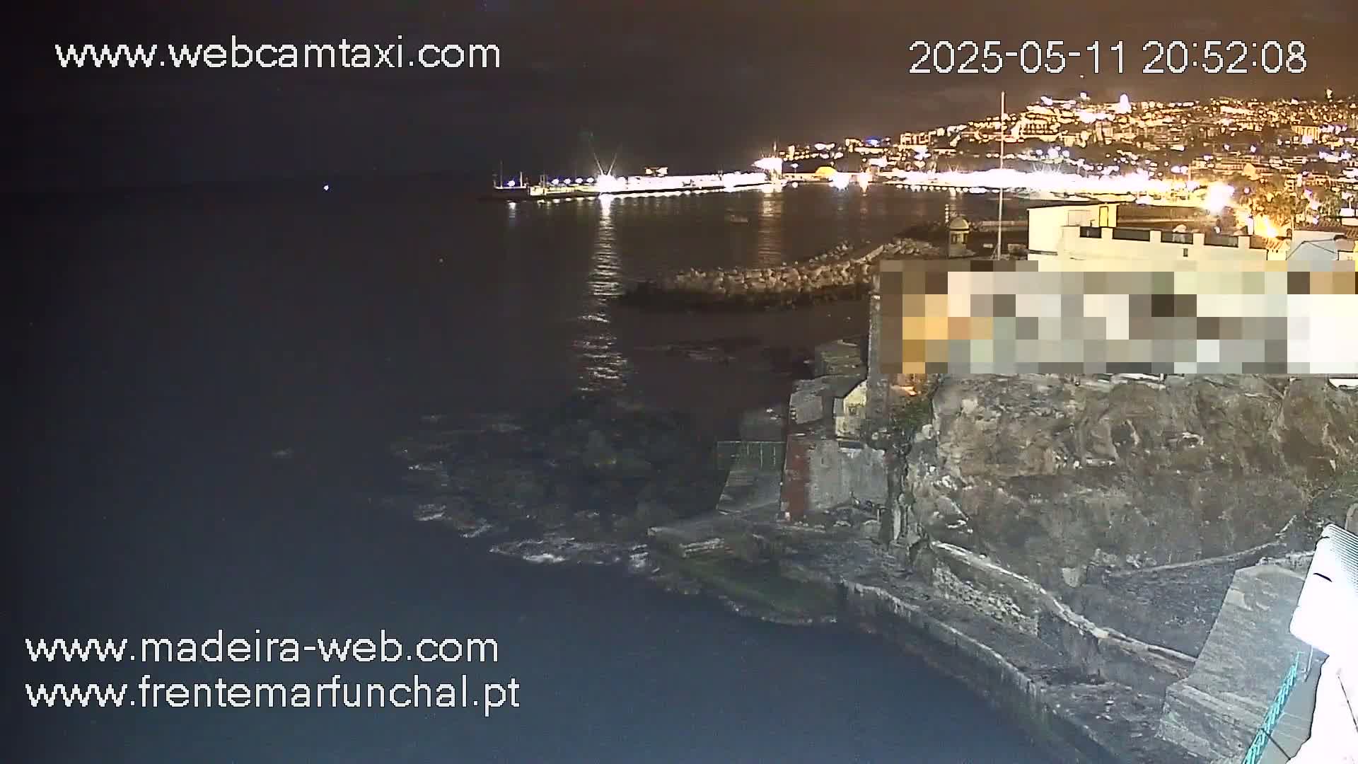 A nighttime view of a rocky coastline, dark water, and a brightly lit city in the distance.