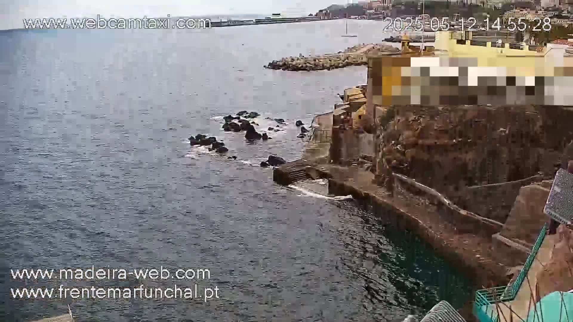 A rocky coastline meets a dark, somewhat choppy sea under an overcast sky, with buildings and structures visible on the land.