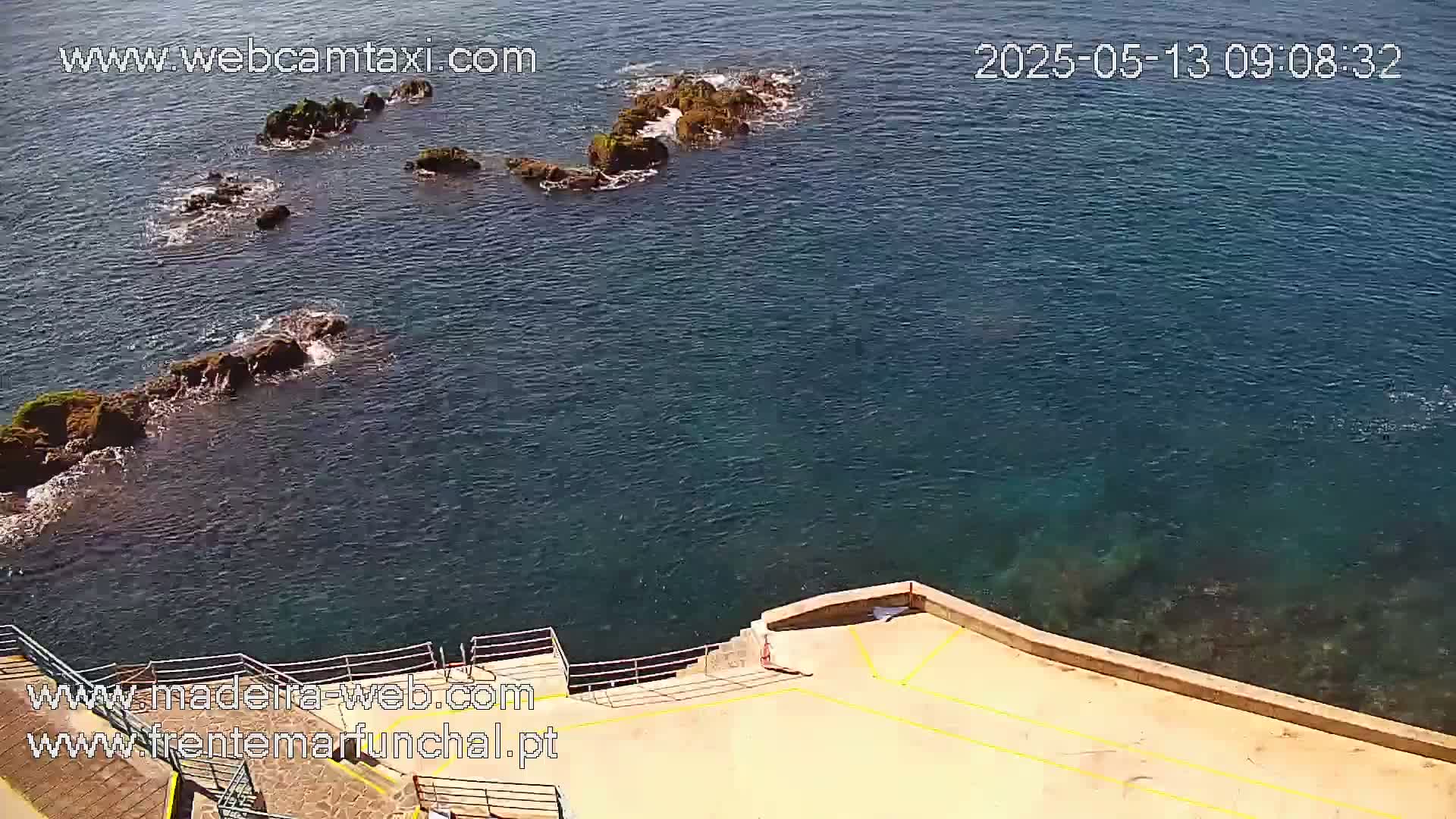 Dark-blue water laps against a rocky shoreline with a light-tan concrete platform and metal railings.