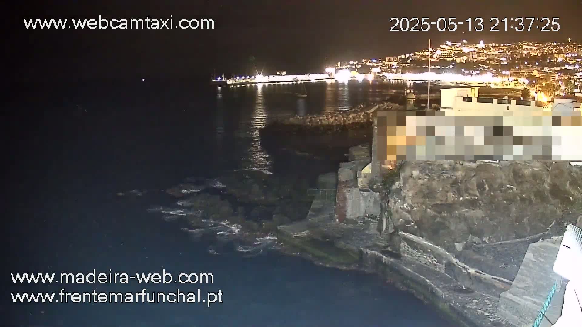 A nighttime coastal scene shows dark water lapping against a rocky shoreline near a city brightly lit in the distance.