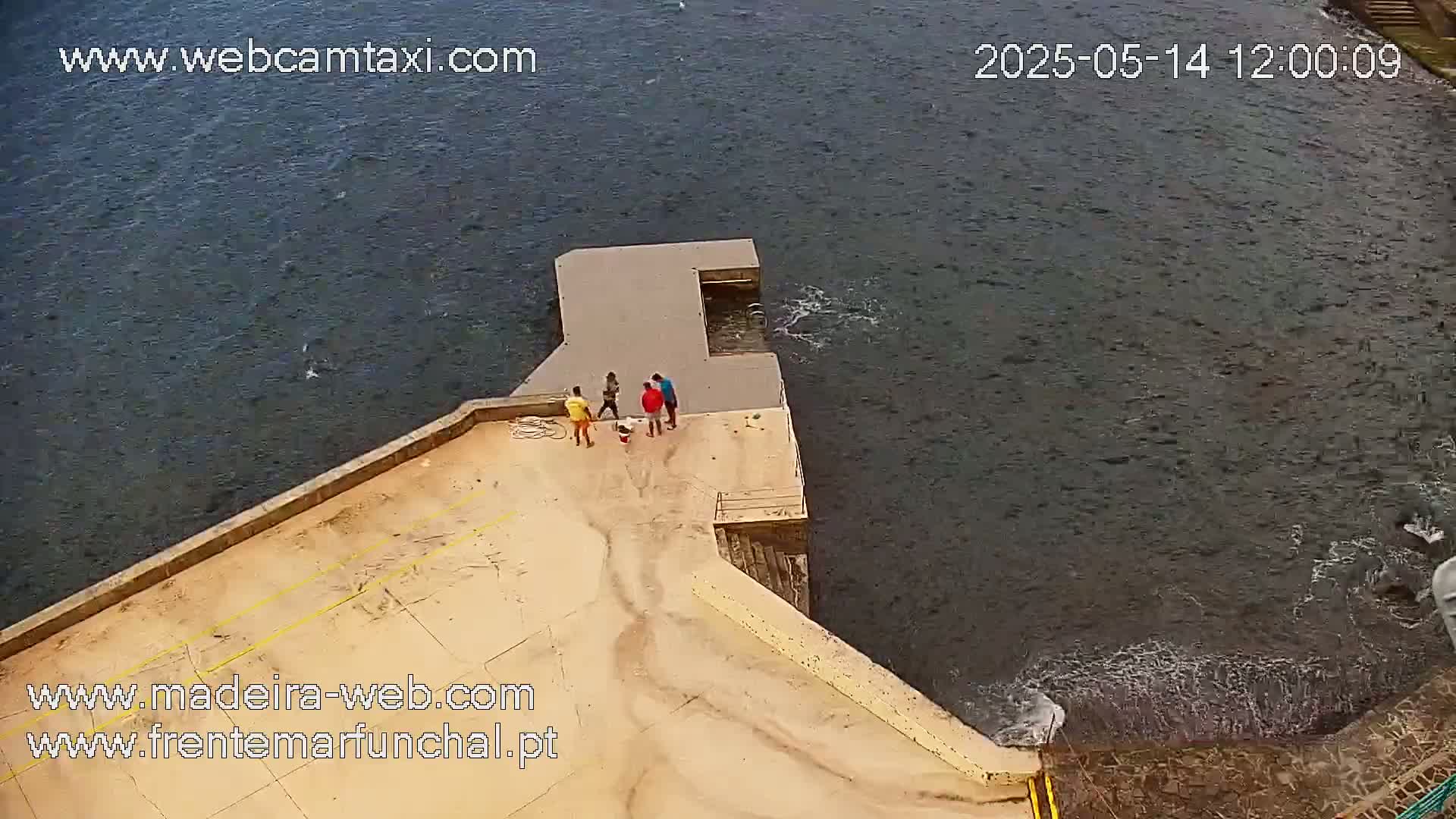A high-angle view shows four people standing on a concrete pier next to choppy dark water.