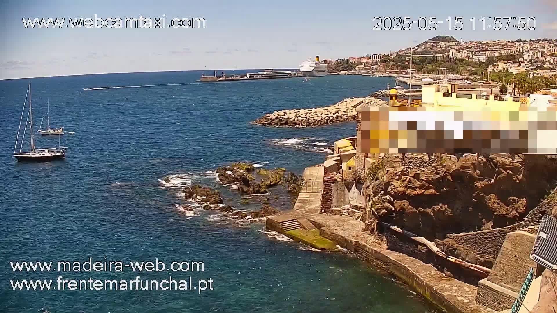 A sunny day shows a coastal view of a harbor with two sailboats, a cruise ship, and rocky coastline with buildings.