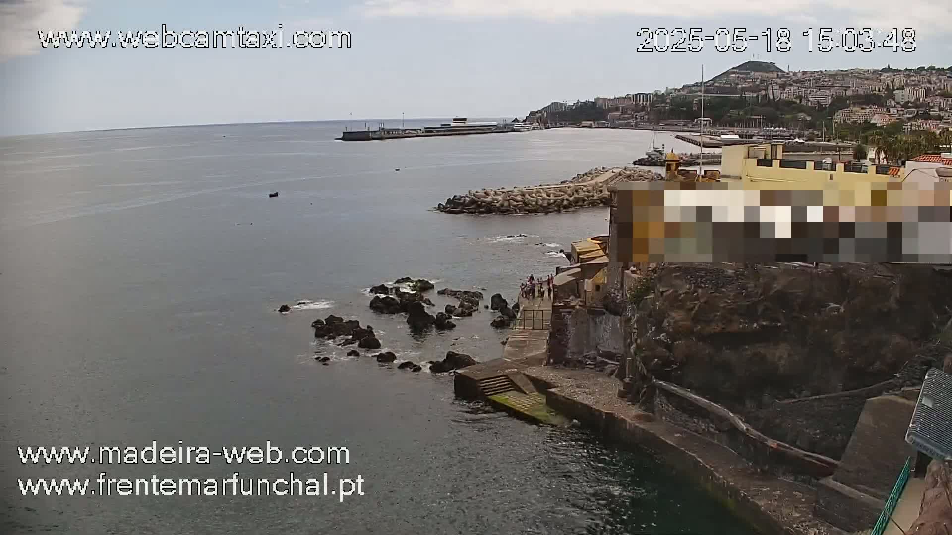 A coastal view on a partly cloudy day shows a rocky shoreline with buildings, a breakwater, and a calm ocean with a town in the background.