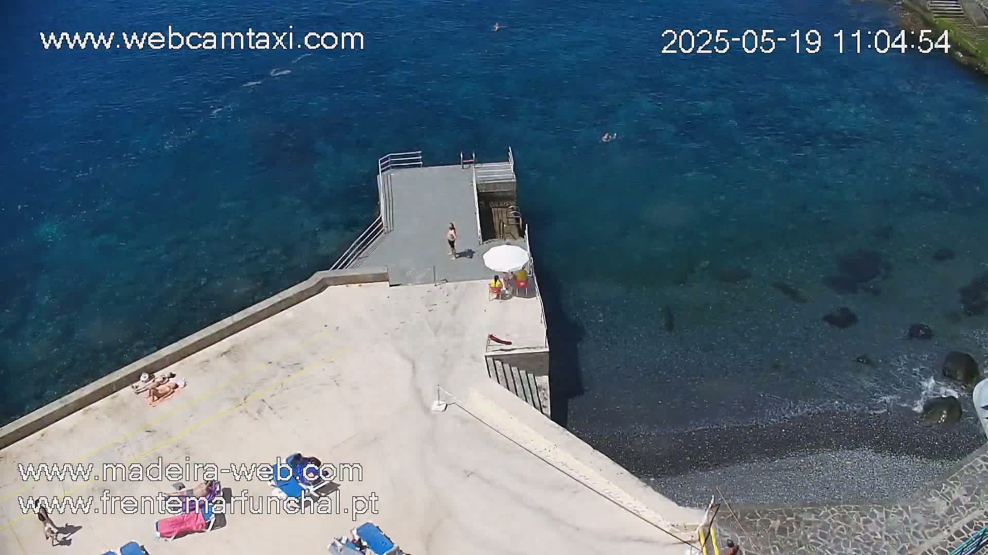 An aerial view shows a small concrete platform and beach area next to clear blue water, with a few people sunbathing and swimming on a sunny day.