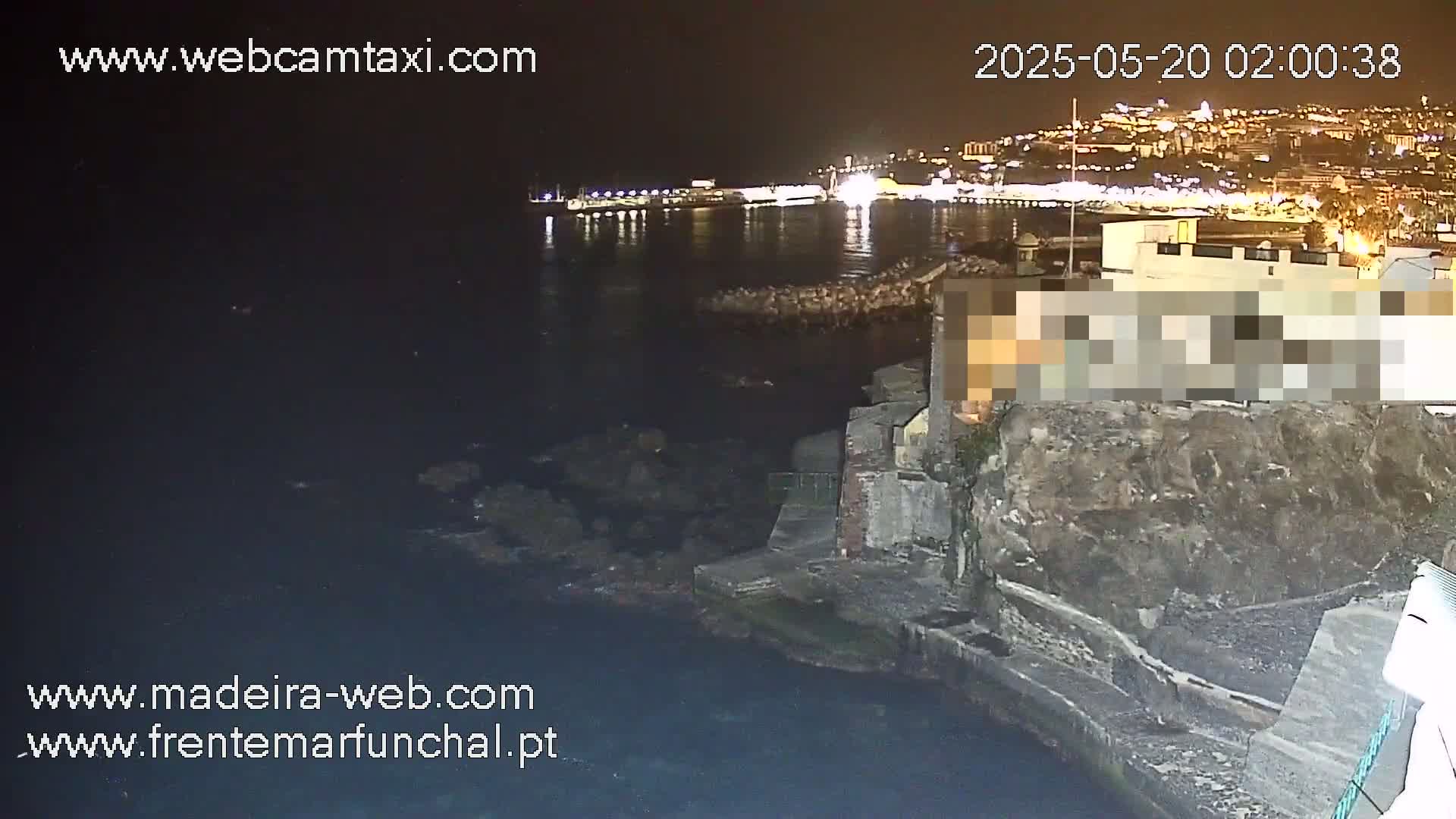 A nighttime coastal scene shows a rocky shoreline with a city's lights illuminating the water in the background.