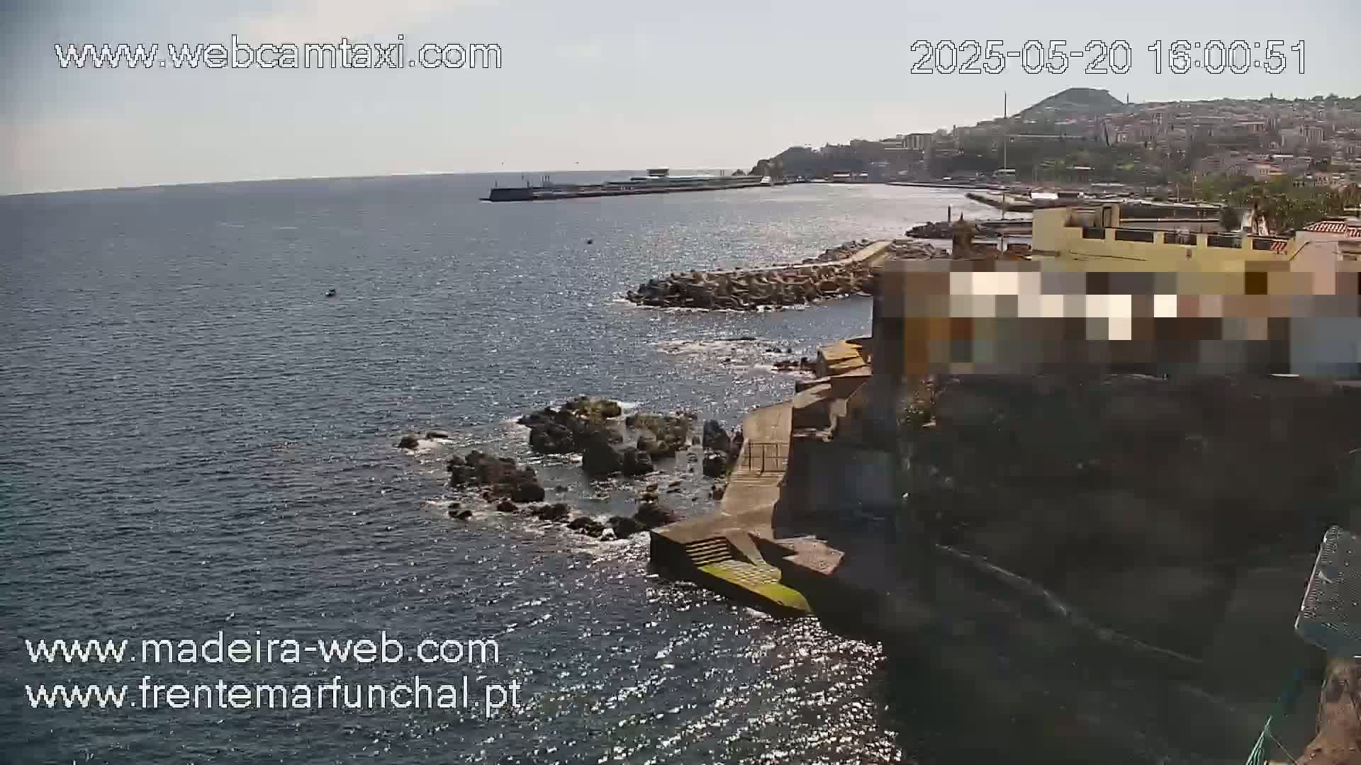 A sunny, seaside view shows dark-blue water lapping against rocky shores and a section of a town built into the coastline.