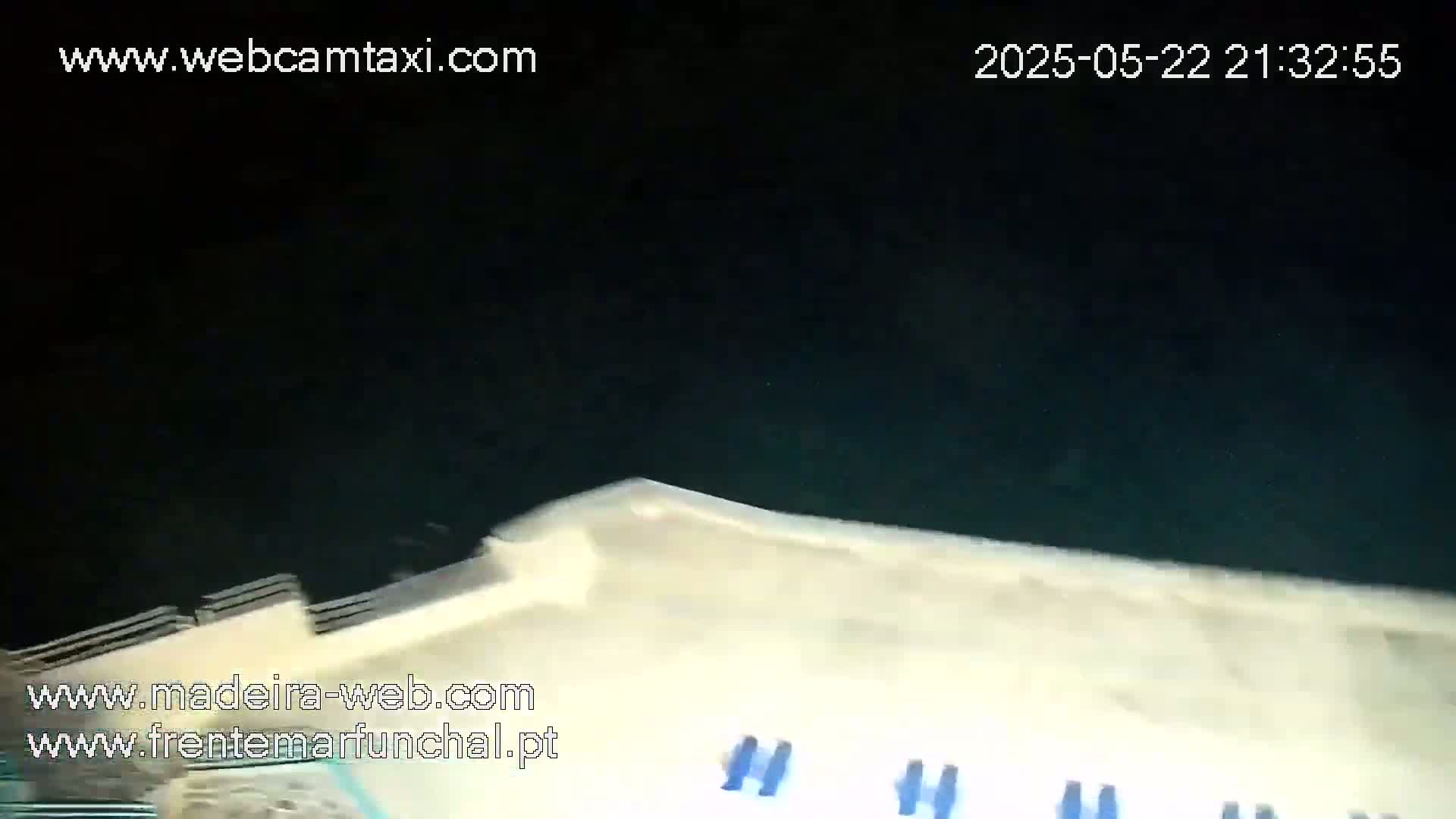 A nighttime, high-angle view shows a sandy beach area next to dark water, with several indistinct objects possibly beach chairs visible on the sand.