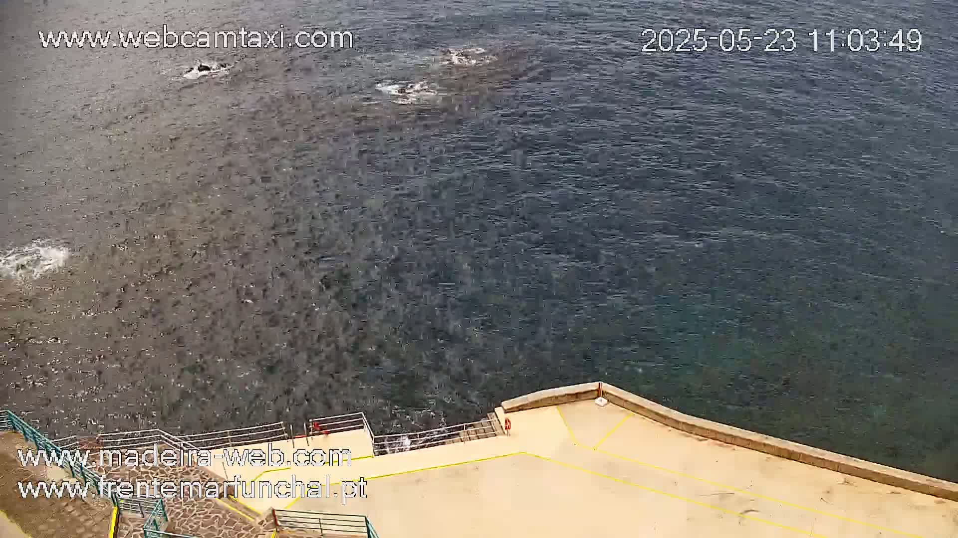 A high-angle view shows dark-blue water with some small waves lapping against a tan concrete area with a yellow line.