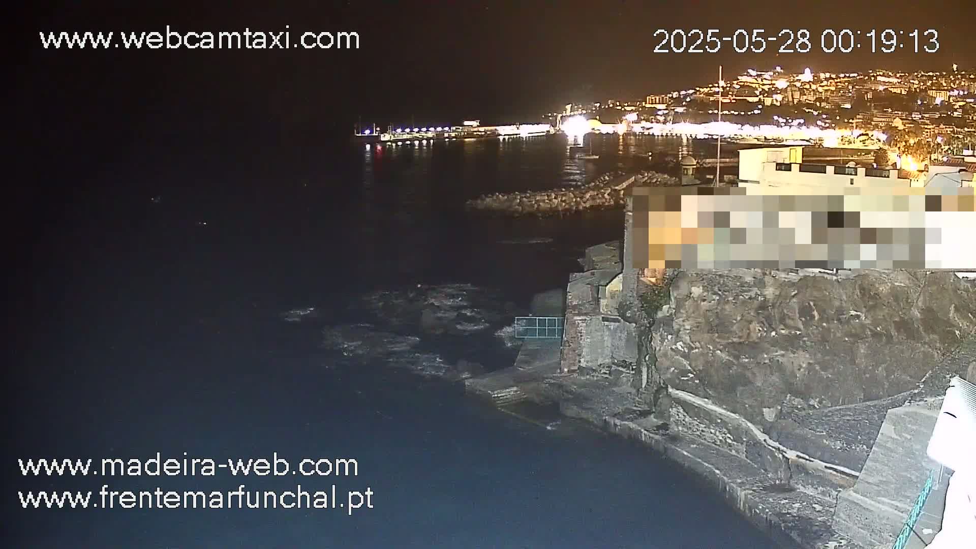 A nighttime coastal scene shows a dark body of water lapping against a rocky shore, with a city's lights twinkling in the distance.