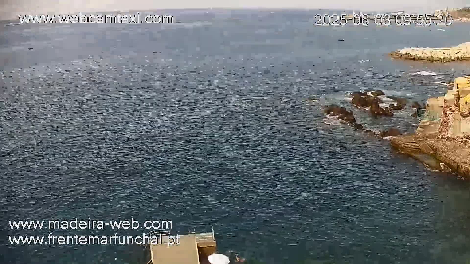 A calm, dark-blue ocean laps against a rocky shoreline, near a stone structure.
