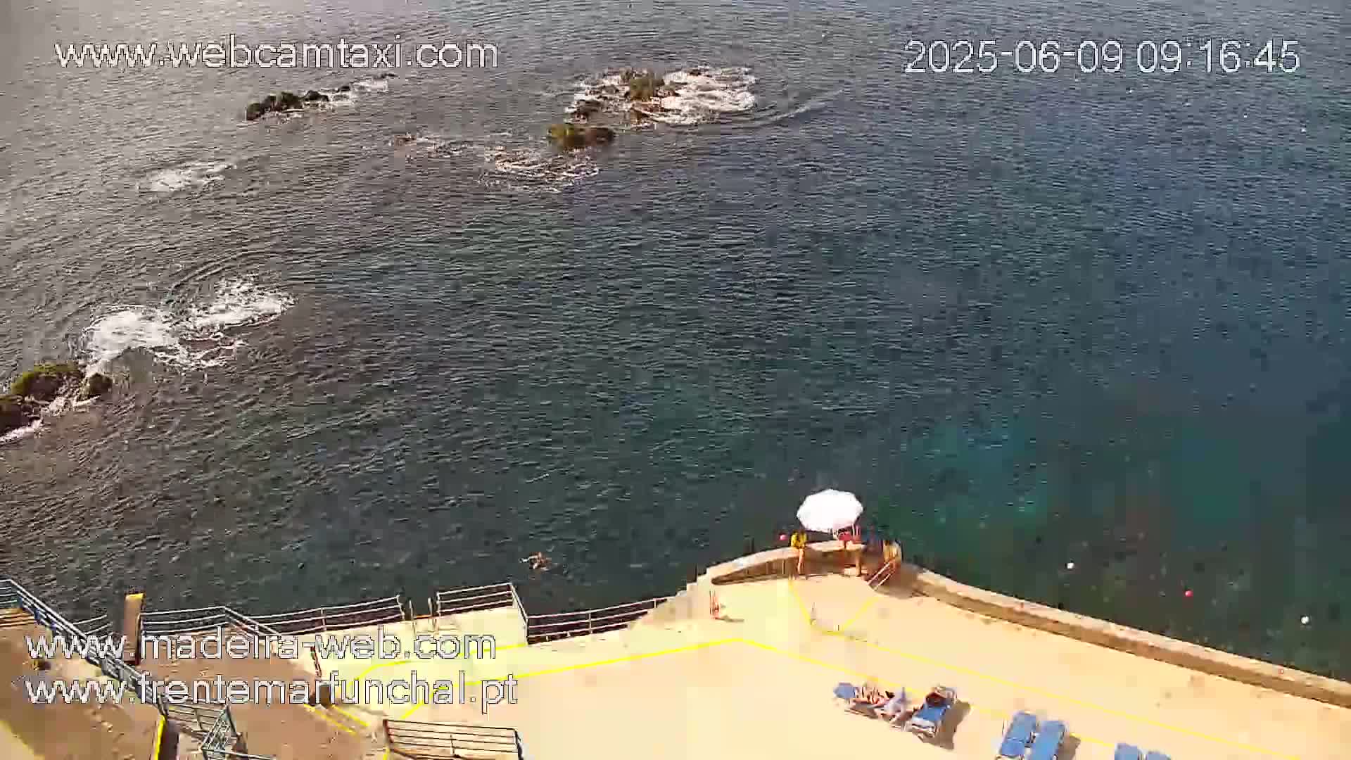 An aerial view shows a rocky coast with calm, dark-blue water, a few people relaxing near a beach area with lounge chairs under an umbrella.