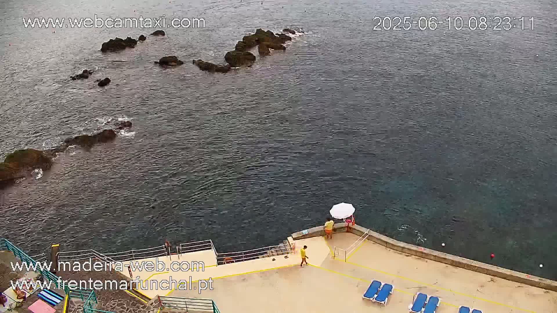 An elevated view shows dark-colored rocks partially submerged in a calm, dark-blue ocean near a tan-colored concrete area with lounge chairs and a few people.