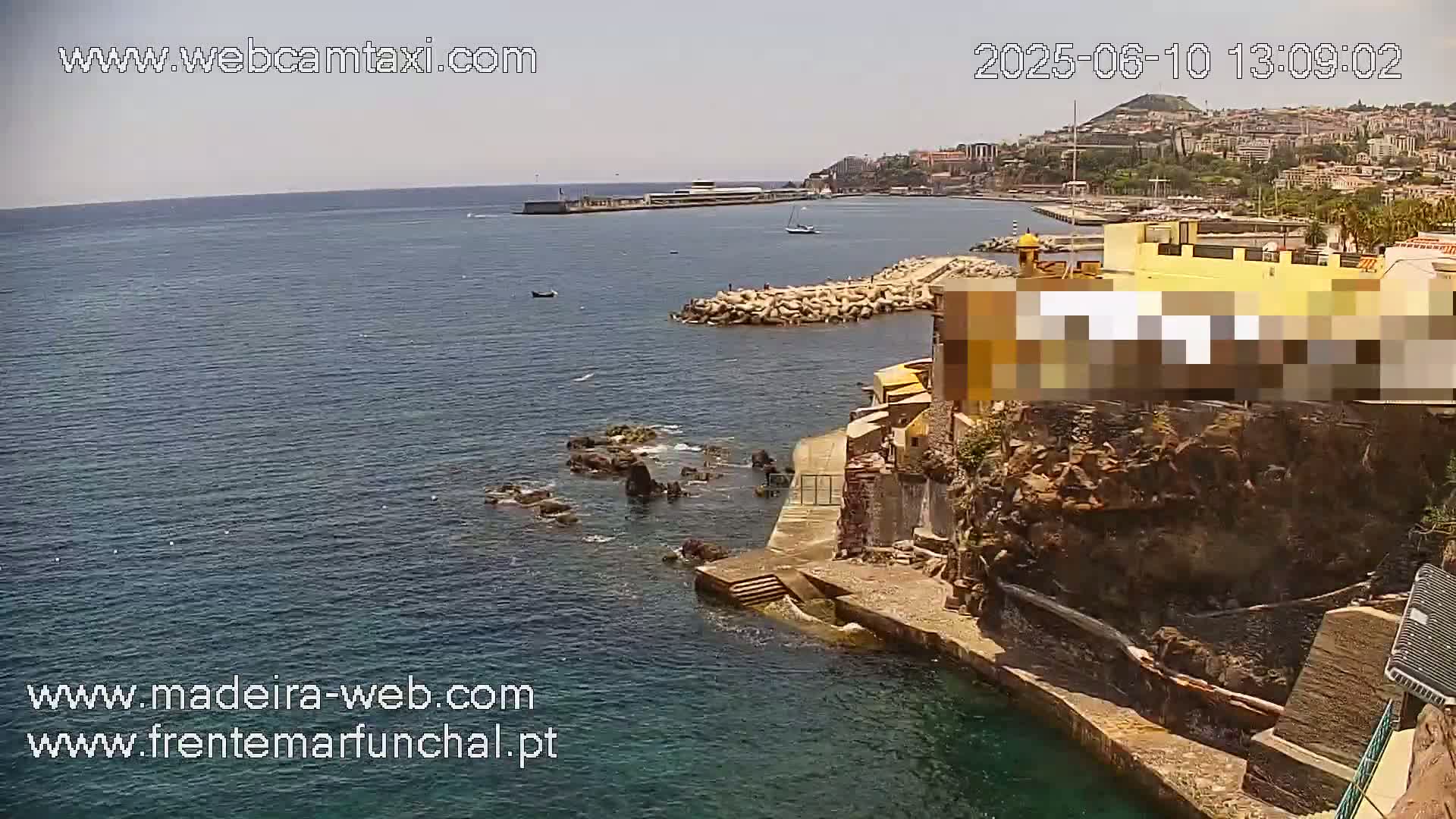A coastal scene on a sunny day shows a rocky shoreline with buildings, a calm ocean, and a distant cityscape.