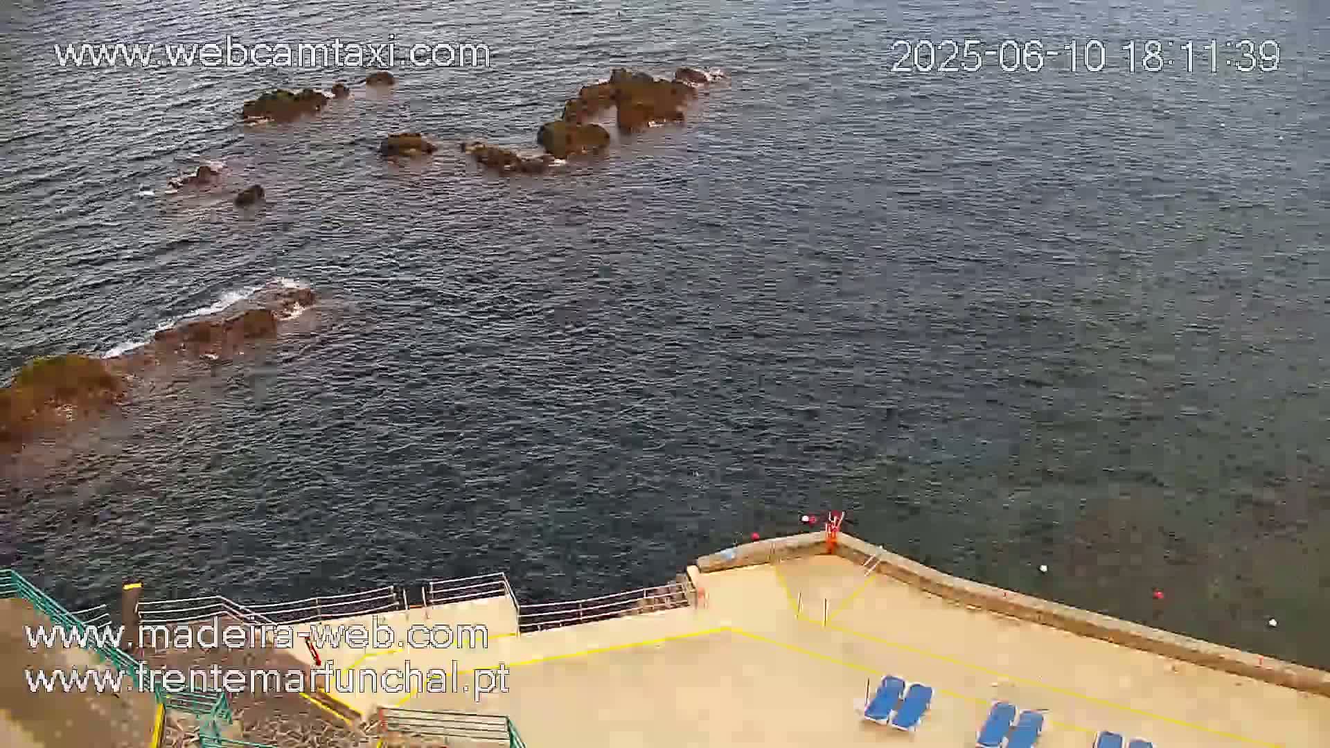 A rocky shoreline meets calm, dark-blue water near a paved area with lounge chairs.