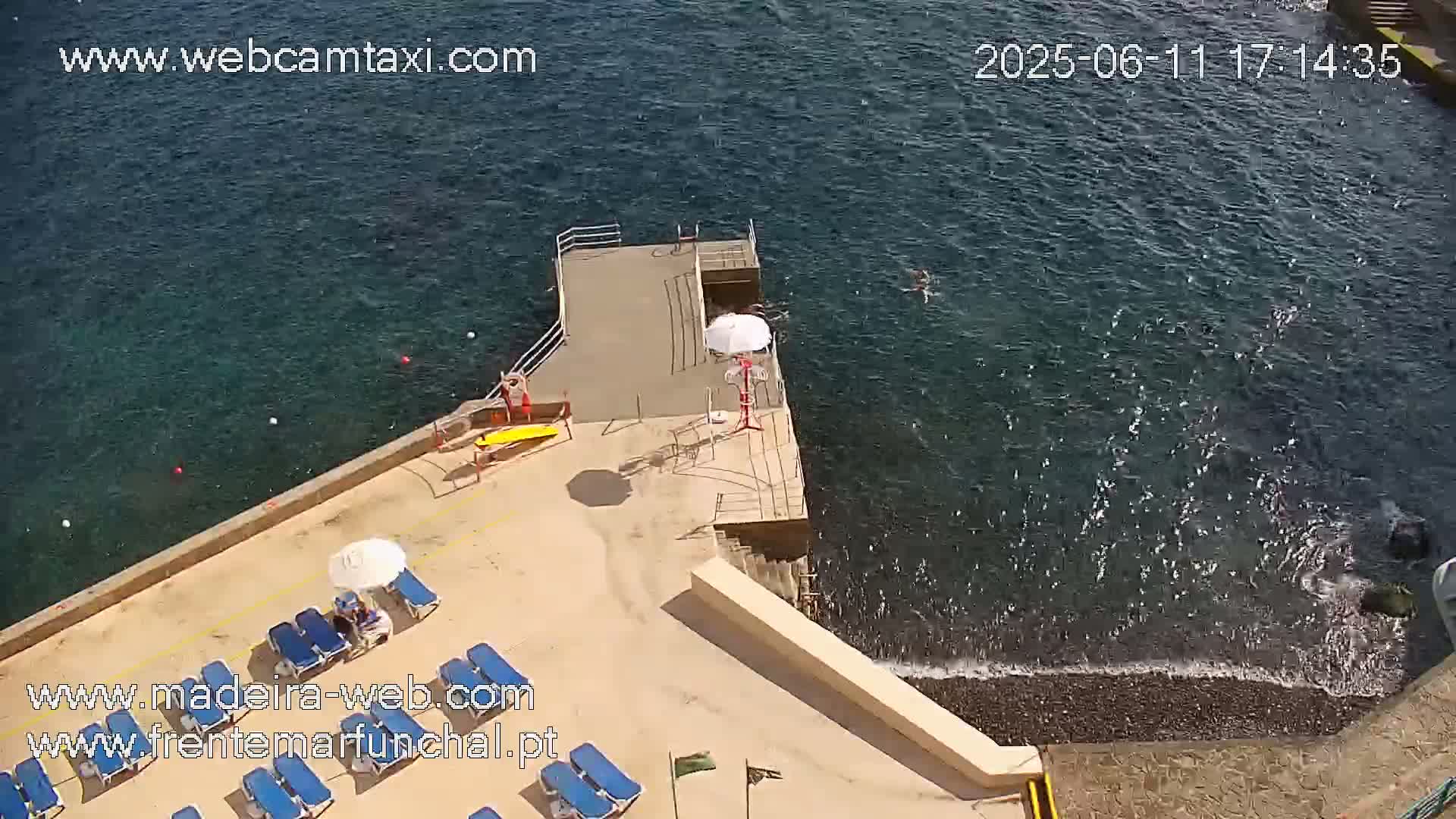 An aerial view shows a beach area with lounge chairs and umbrellas, a small dock extending into dark-blue water with small waves lapping a pebble shore, and several people swimming.