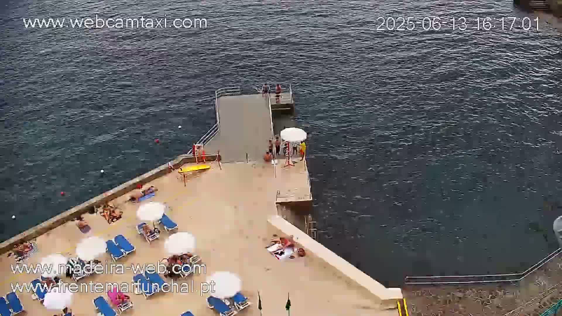An aerial view shows a small beach with sunbathers and several umbrellas near a short pier extending into dark, somewhat choppy water.