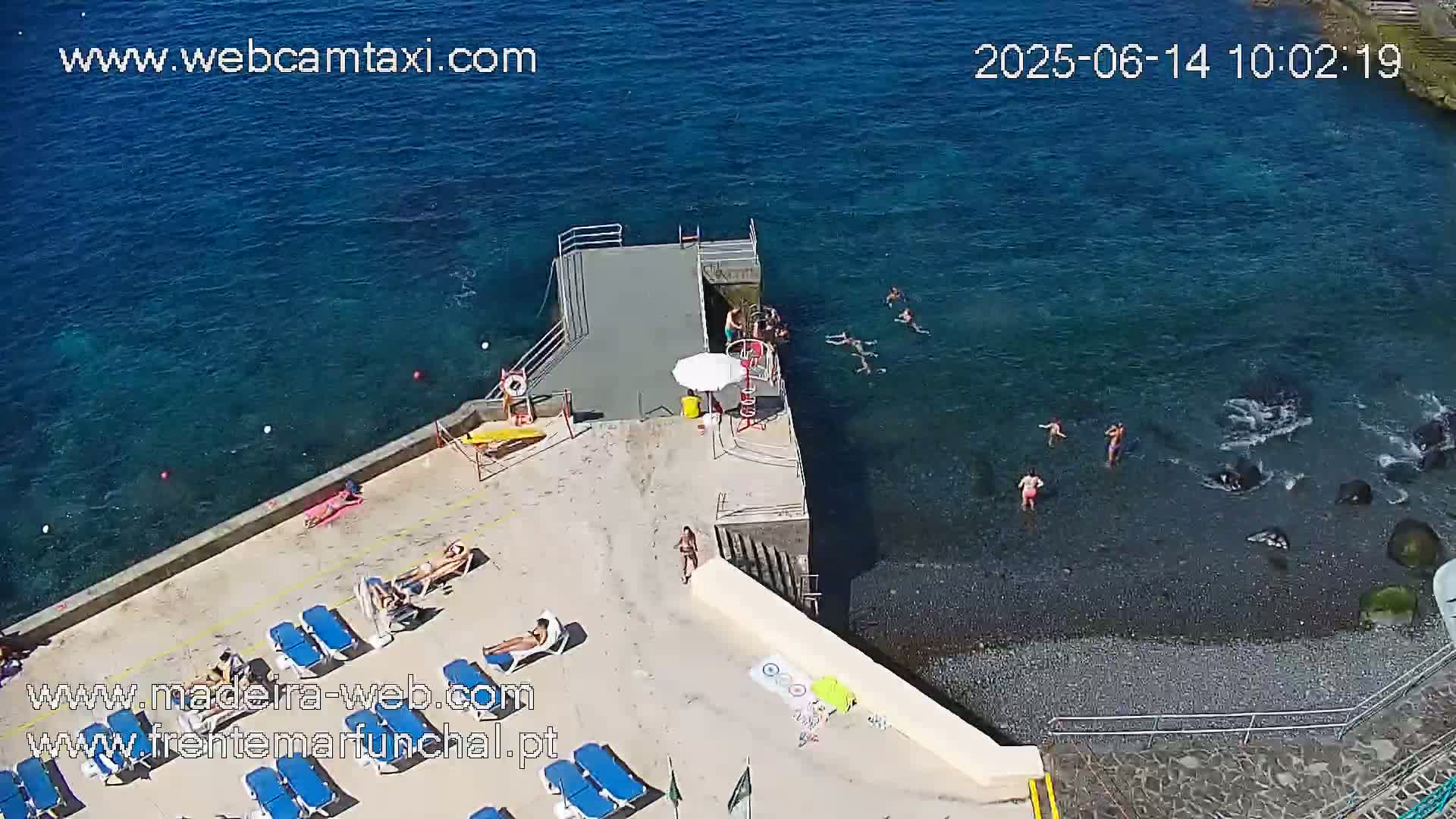 An aerial view shows a small beach with people swimming in clear blue water, sunbathing on lounge chairs, and standing on a concrete platform next to a dock.