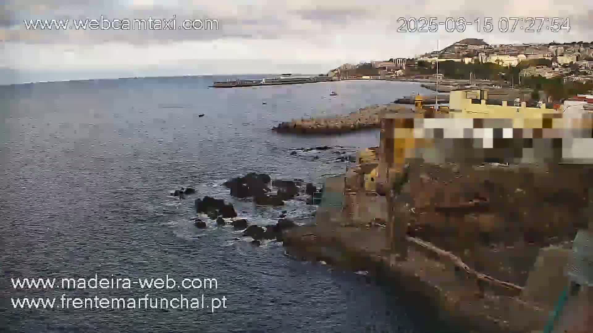 A coastal view shows dark-colored water with dark rocks and a building on a cliff overlooking a calm ocean under an overcast sky.