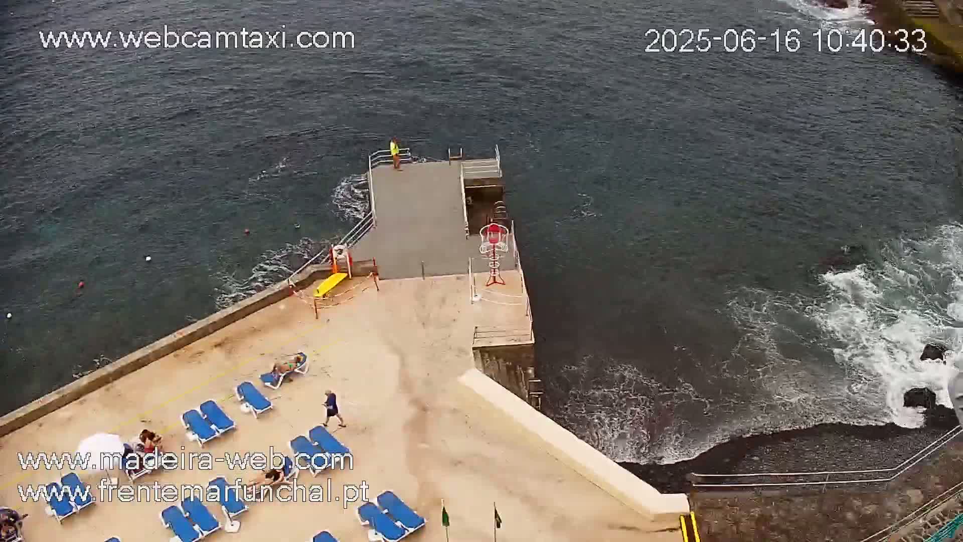A sunbathers lounge on a patio overlooking a dark-blue ocean with small waves crashing against a rocky shore.