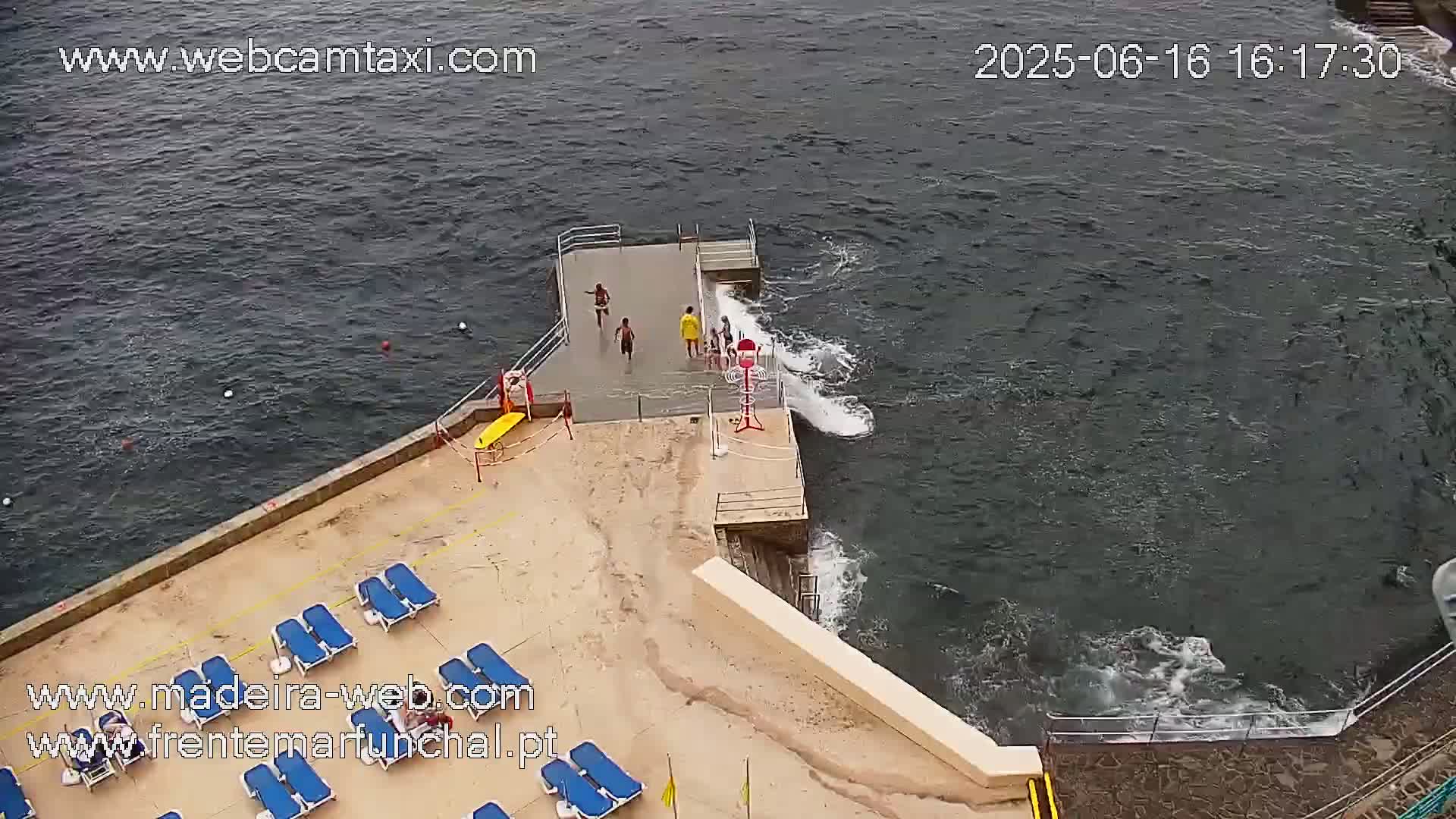 An aerial view shows a small, rocky beach area with several people near a short pier extending into dark, somewhat choppy water.