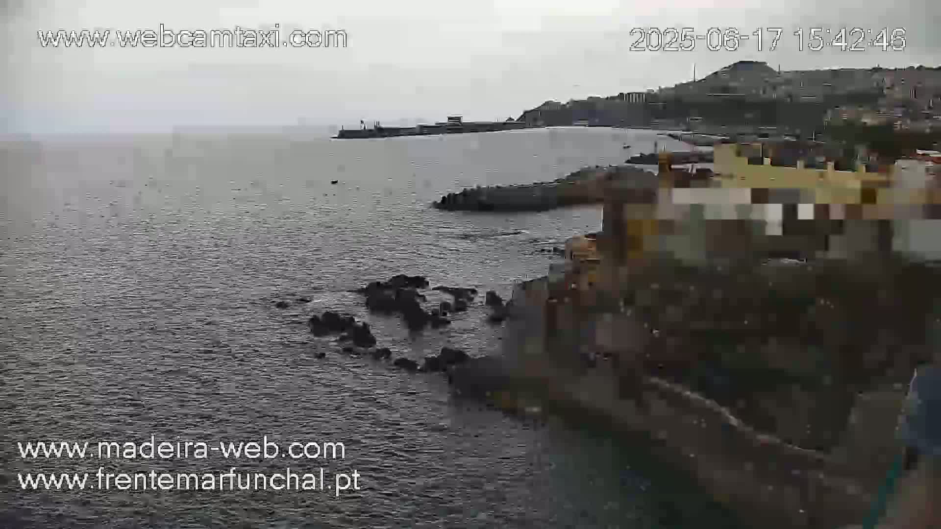 A dark, overcast day shows a rocky coastline and calm ocean water, with a town visible in the distance.