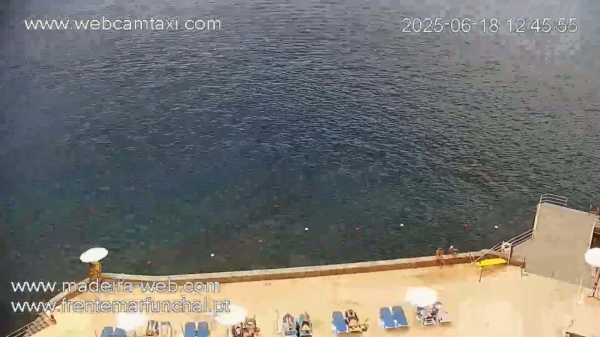 A calm body of water is seen from an elevated view, next to a sandy area with lounge chairs and people relaxing, on a sunny day.