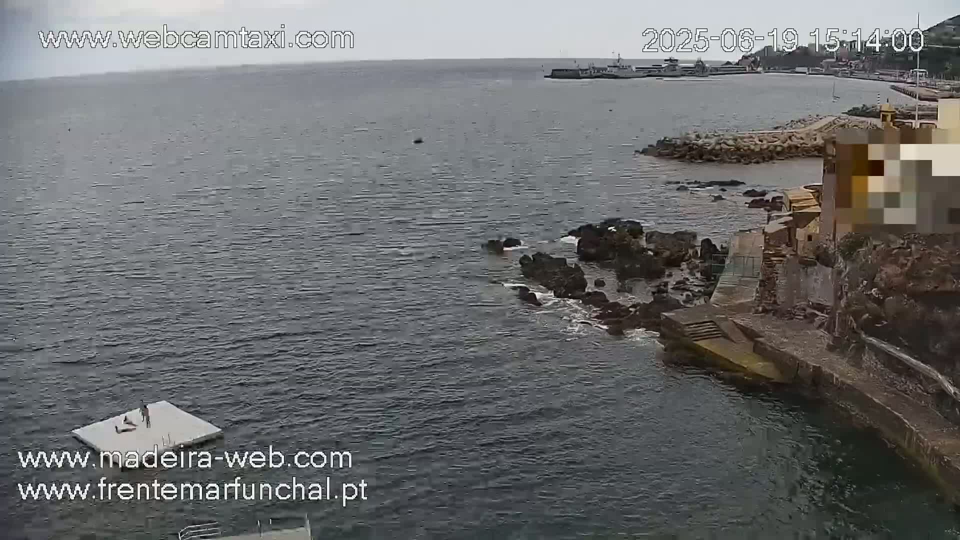 A mostly cloudy day reveals a rocky coast with a small,  man-made platform in the water, and a distant harbor; a building is visible along the shore.