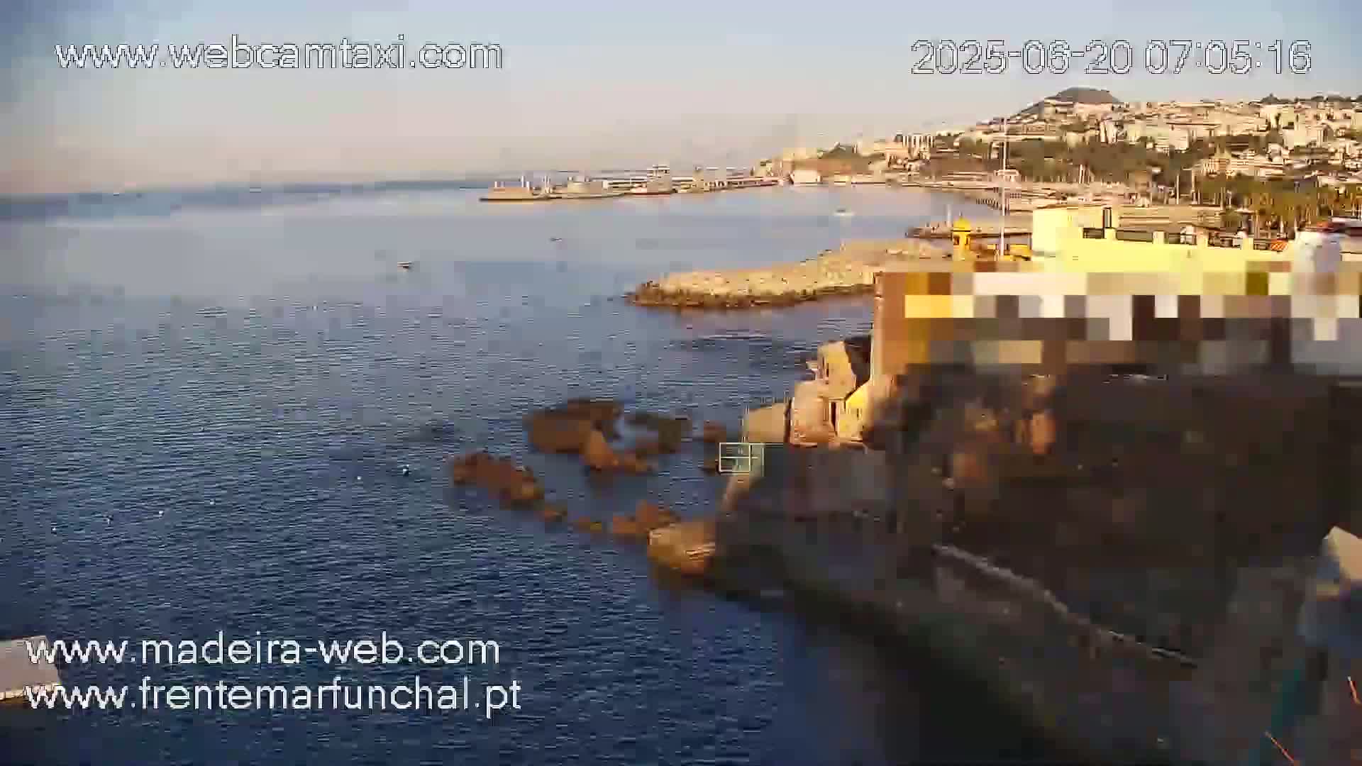 A coastal scene on a sunny morning shows a calm ocean with dark rocks near a shoreline, a town visible in the background.