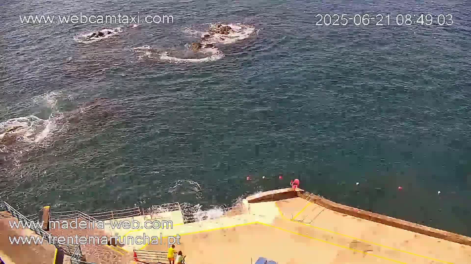A rocky outcrop in a moderately choppy ocean is visible from an elevated vantage point overlooking a paved area near the water's edge.