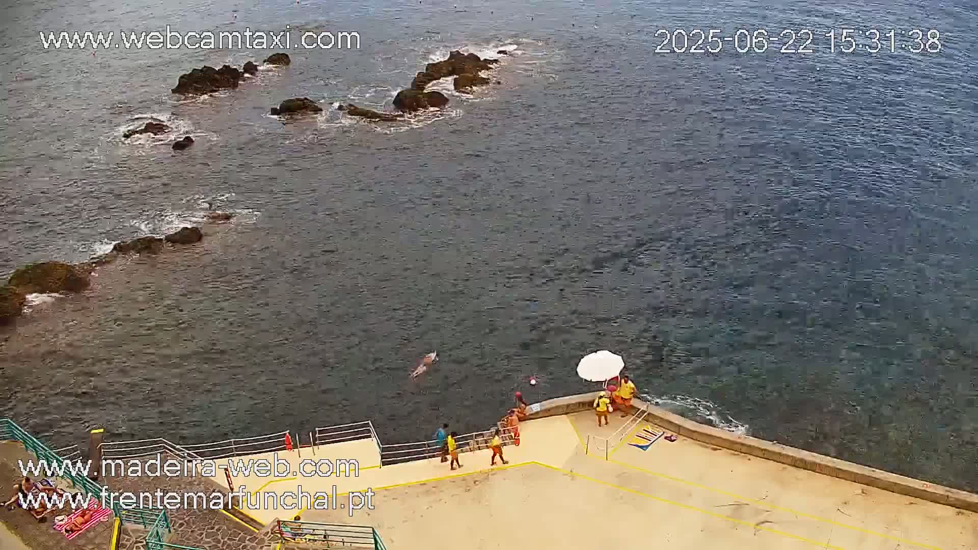 A high-angle view shows a rocky coastline with calm, dark-blue water, a few people on a concrete platform near the water's edge, and one person swimming in the sea.