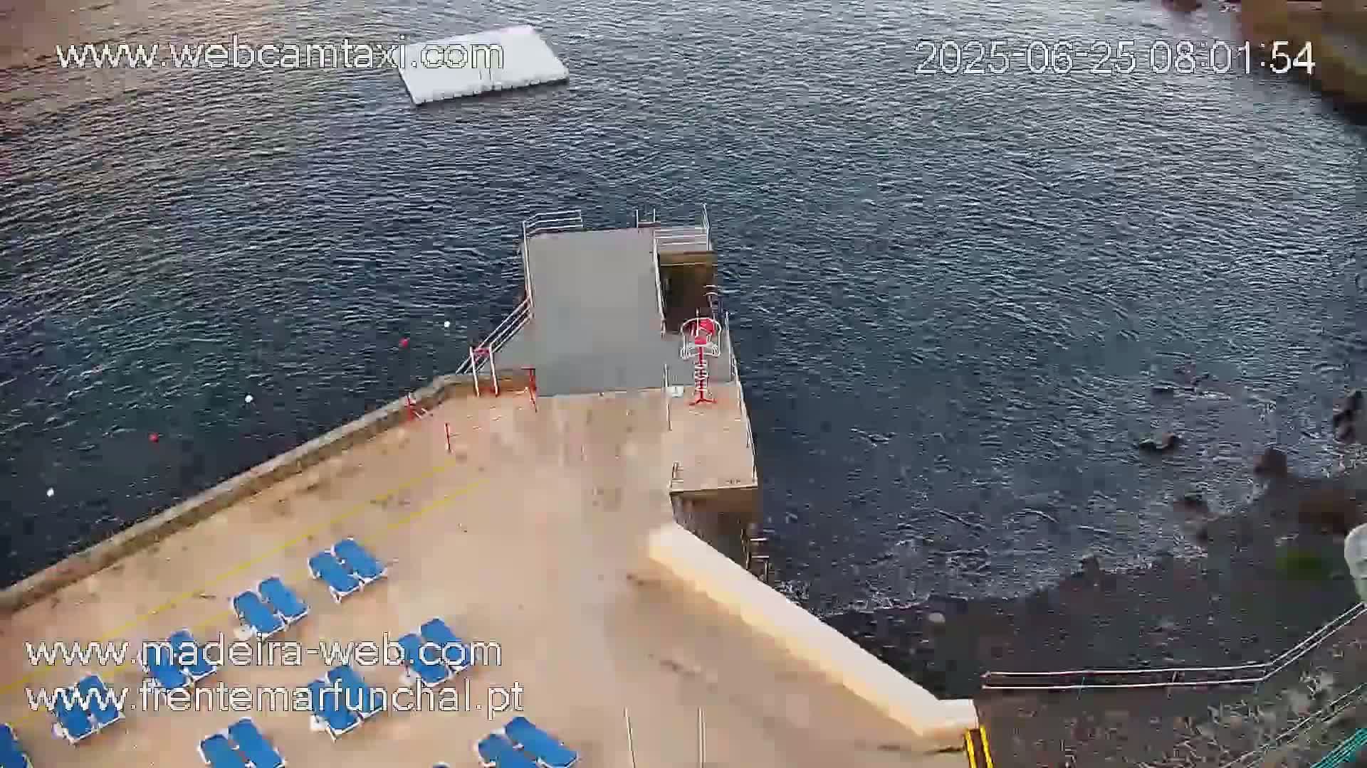 An aerial view shows a concrete deck with lounge chairs next to dark, choppy water, a floating platform, and a small diving platform.