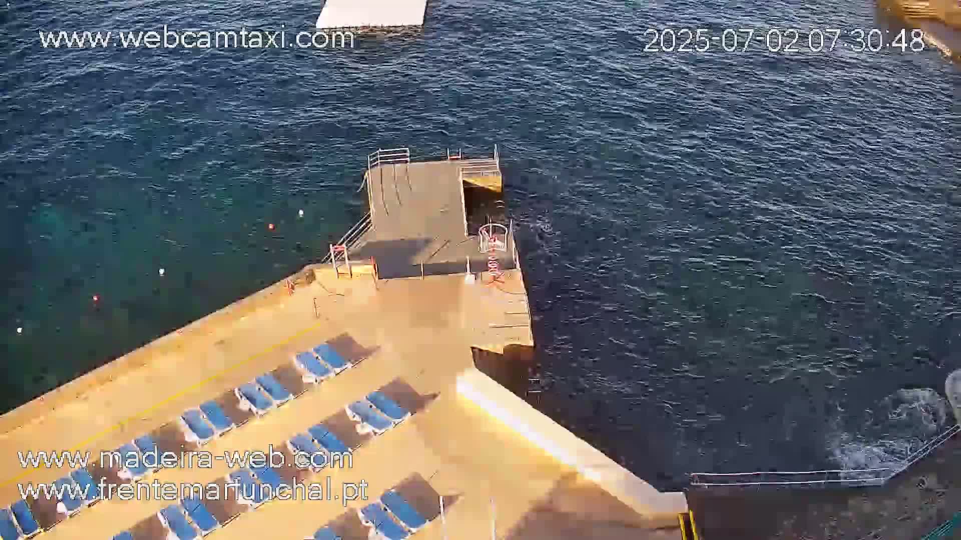 An aerial view shows a mostly empty sunbathing area with a small dock extending into dark blue water.
