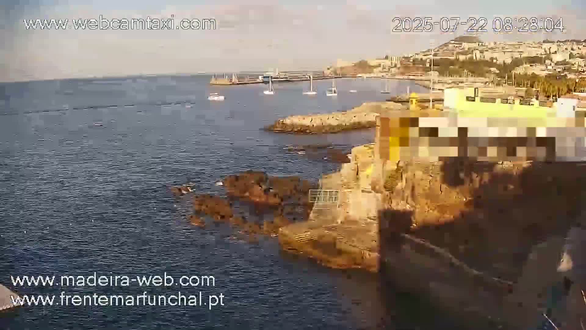 A partly sunny day shows a rocky coast with a calm body of water containing several small boats and a distant cityscape in the background.